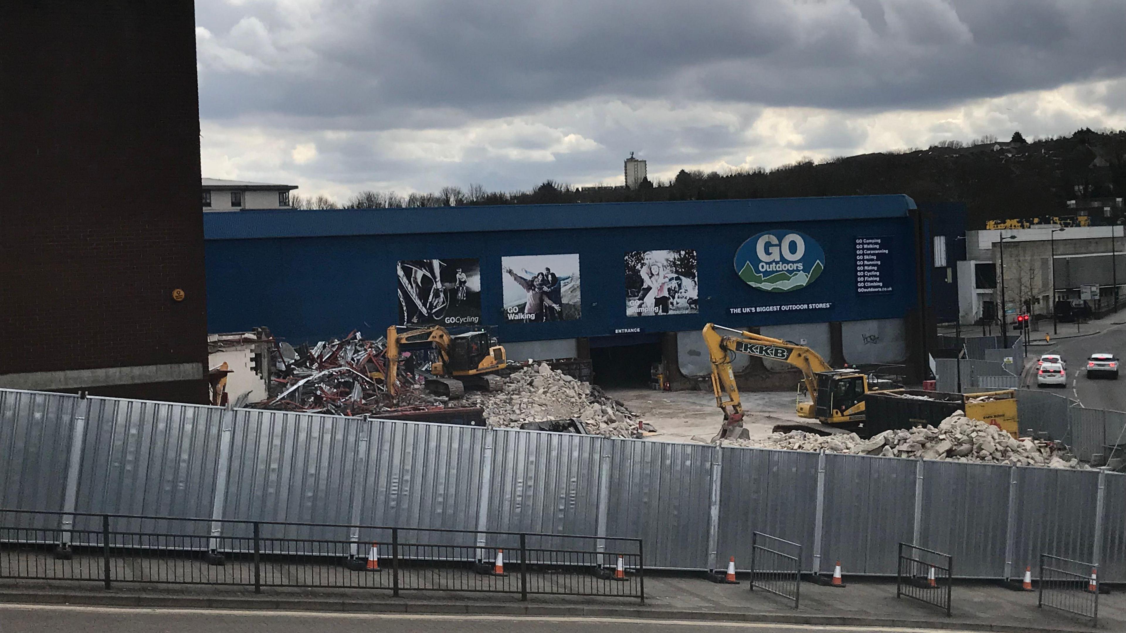 Two diggers tearing down a former retail site surrounded by rubble. The remaining building is blue with a Go Outdoors logo on the side.
