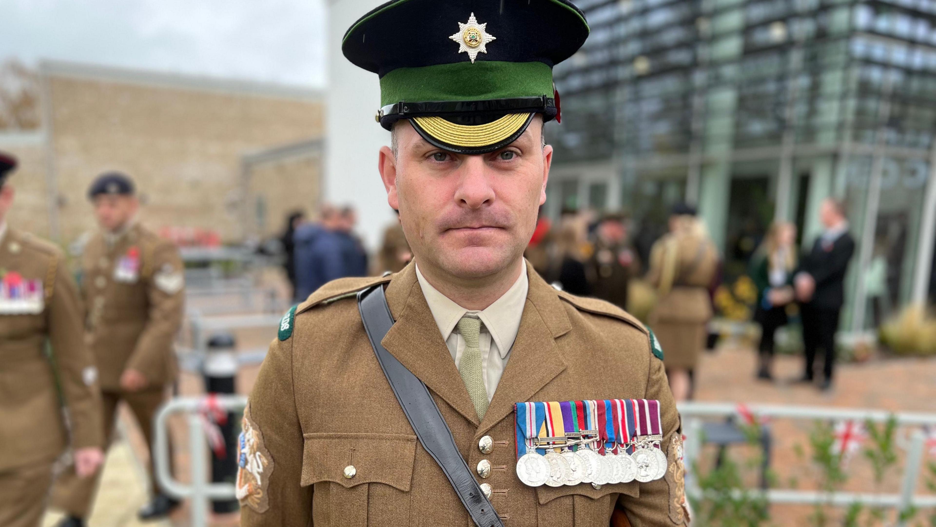 Garrison Sergeant Major K Tomlinson. He is wearing a brown military uniform and a black hat. There are several medals pinned to his jacket. He is looking at the camera with a blank expression.