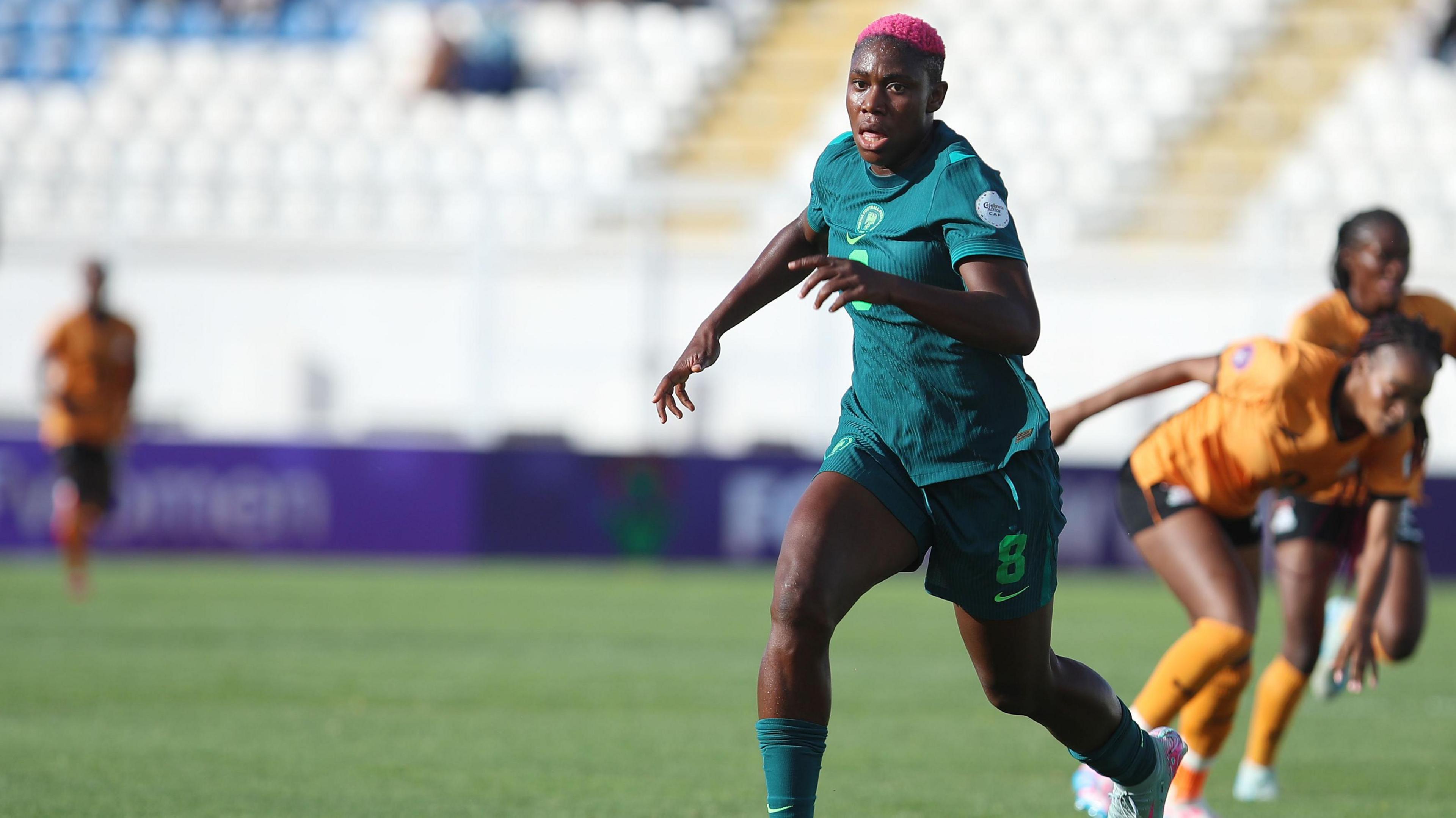 Asisat Oshoala, wearing an all-green Nigeria kit with number 8 written on it, chases a ball during a match against Zambia, with three opponents in copper jerseys and socks and black shorts seen in the background on the green pitch. An empty stand with white seats is blurred in the background