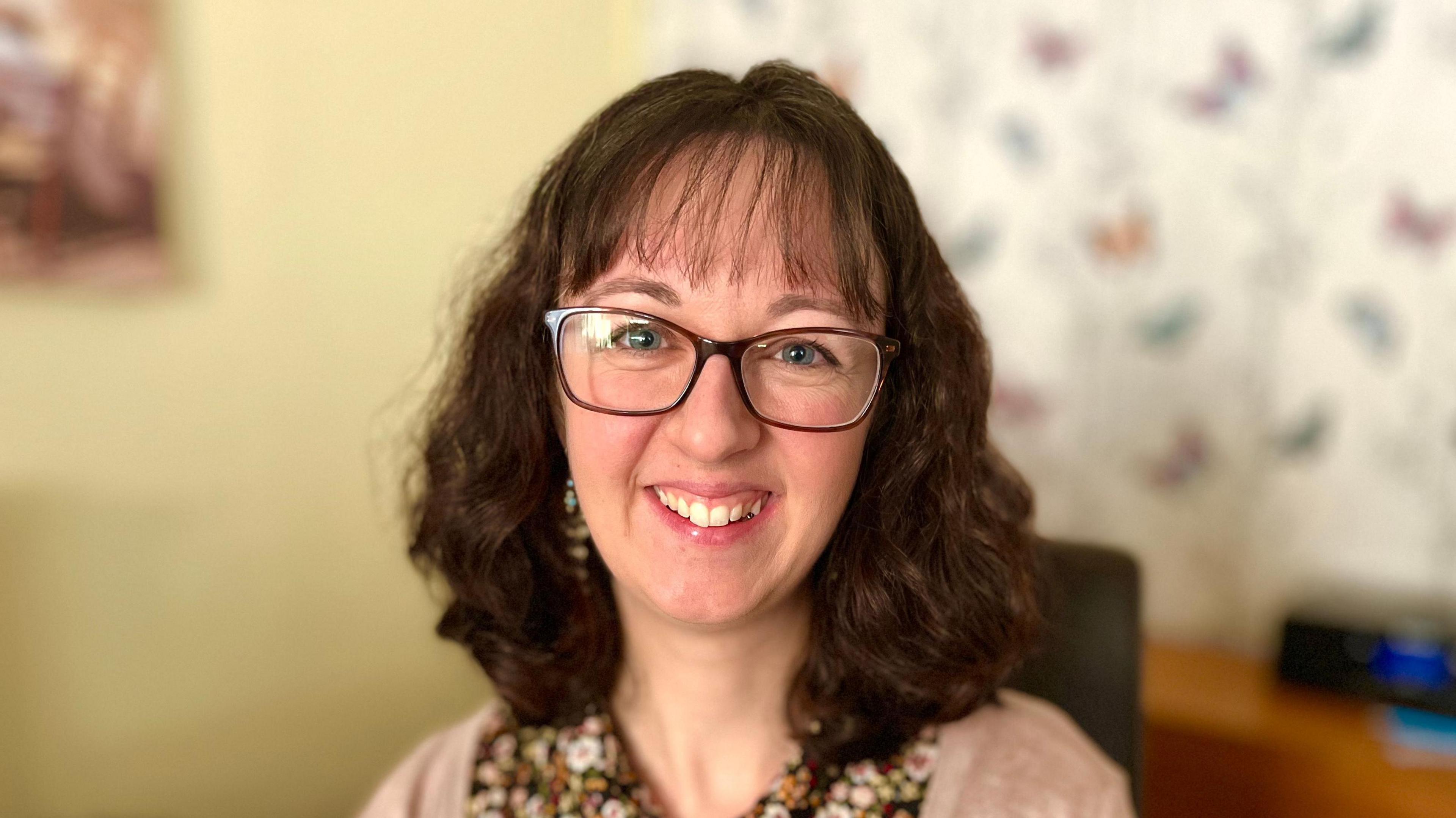 Woman with wavy brown hair smiling to camera. She had a cardigan on over a floral top. 