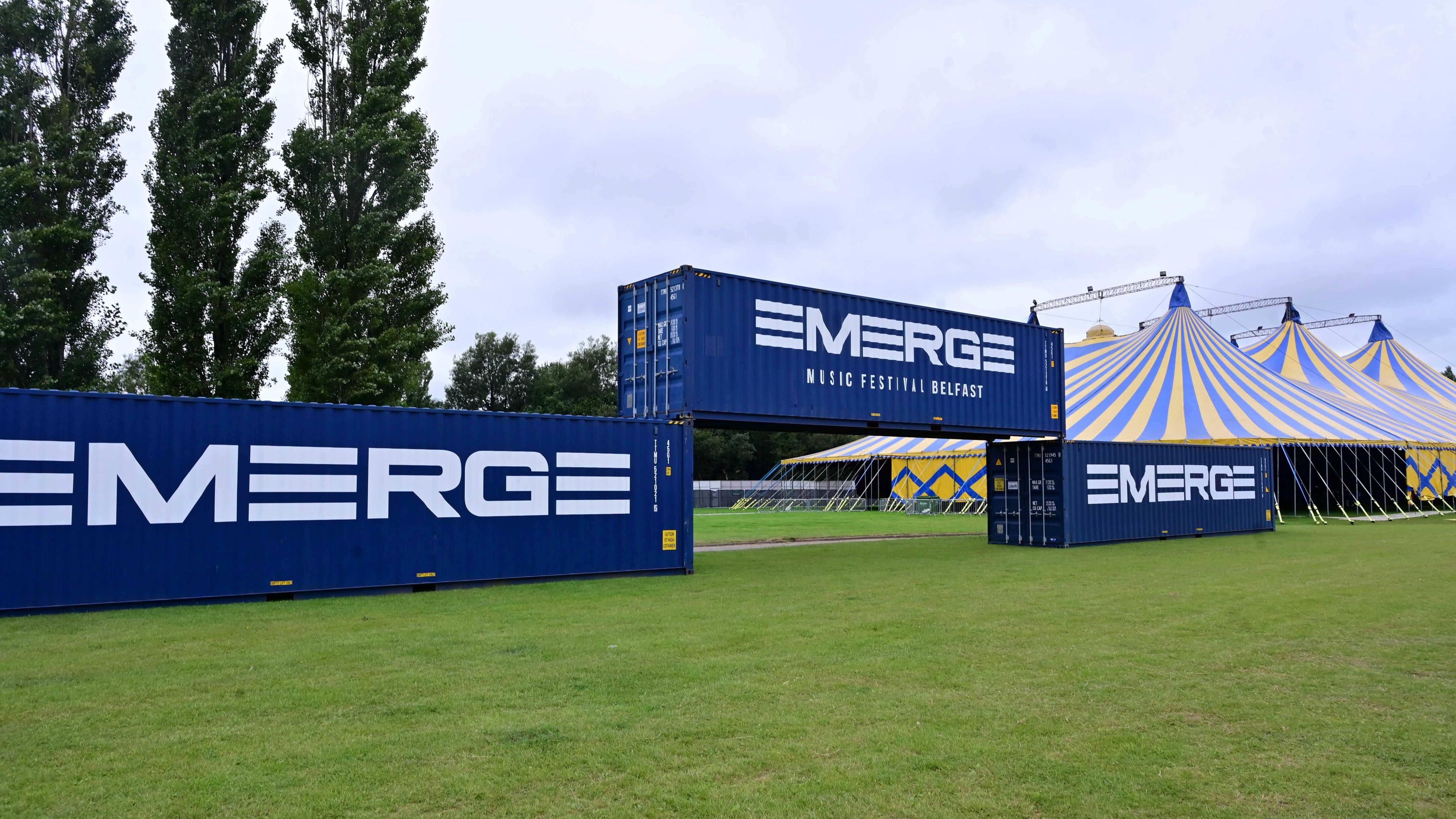 Three large blue containers. The container in the middle is resting on top of either end of the other two containers. Each container has the word 'EMERGE' written in white on it. Behind the containers are a number of trees, as well as some large blue and yellow tents.