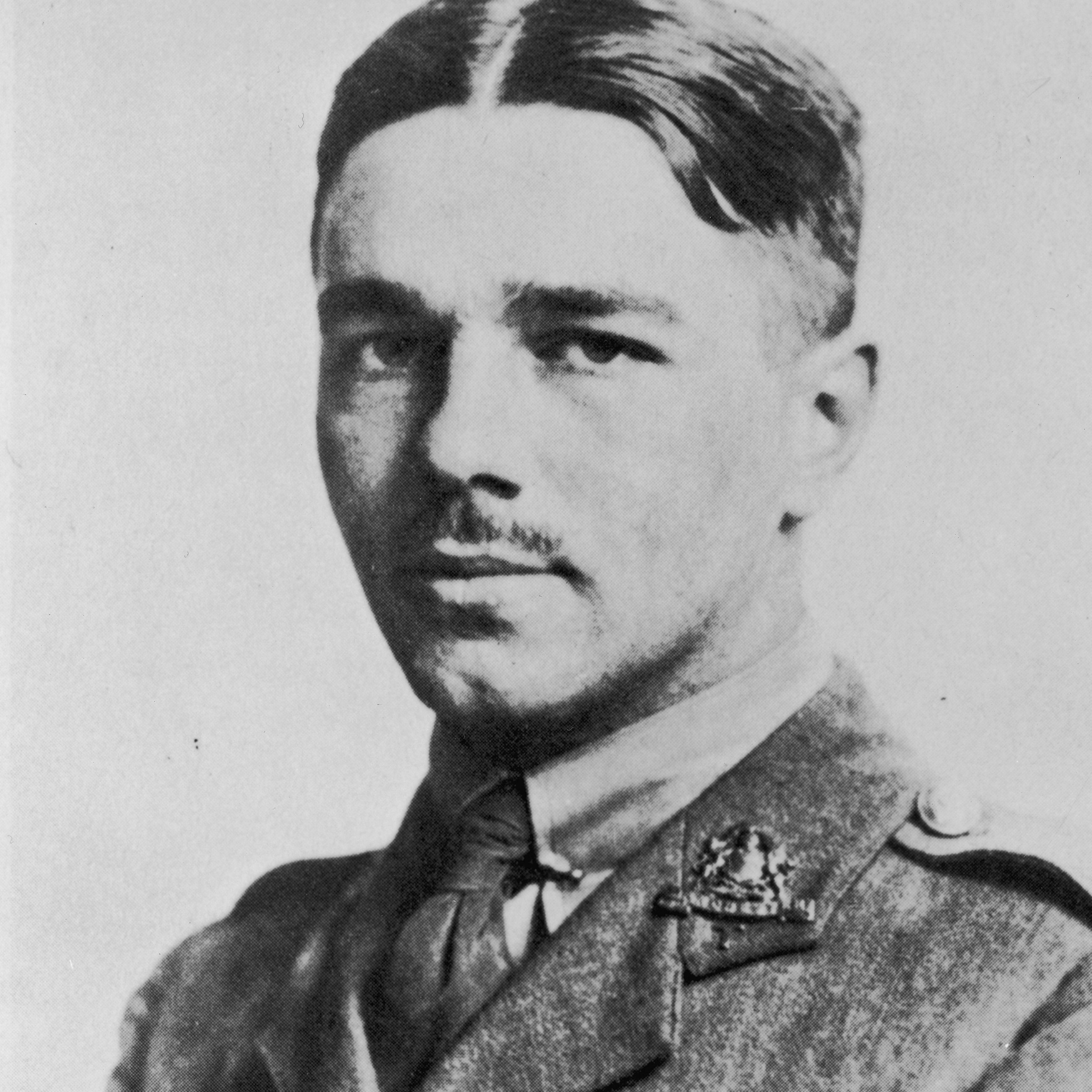 A formal black and white portrait of Officer Cadet Wilfred Owen, circa 1916, shows him wearing a shirt, jacket and tie, with a badge on his lapel. He has a moustache and his hair is parted in the middle and smoothed down.