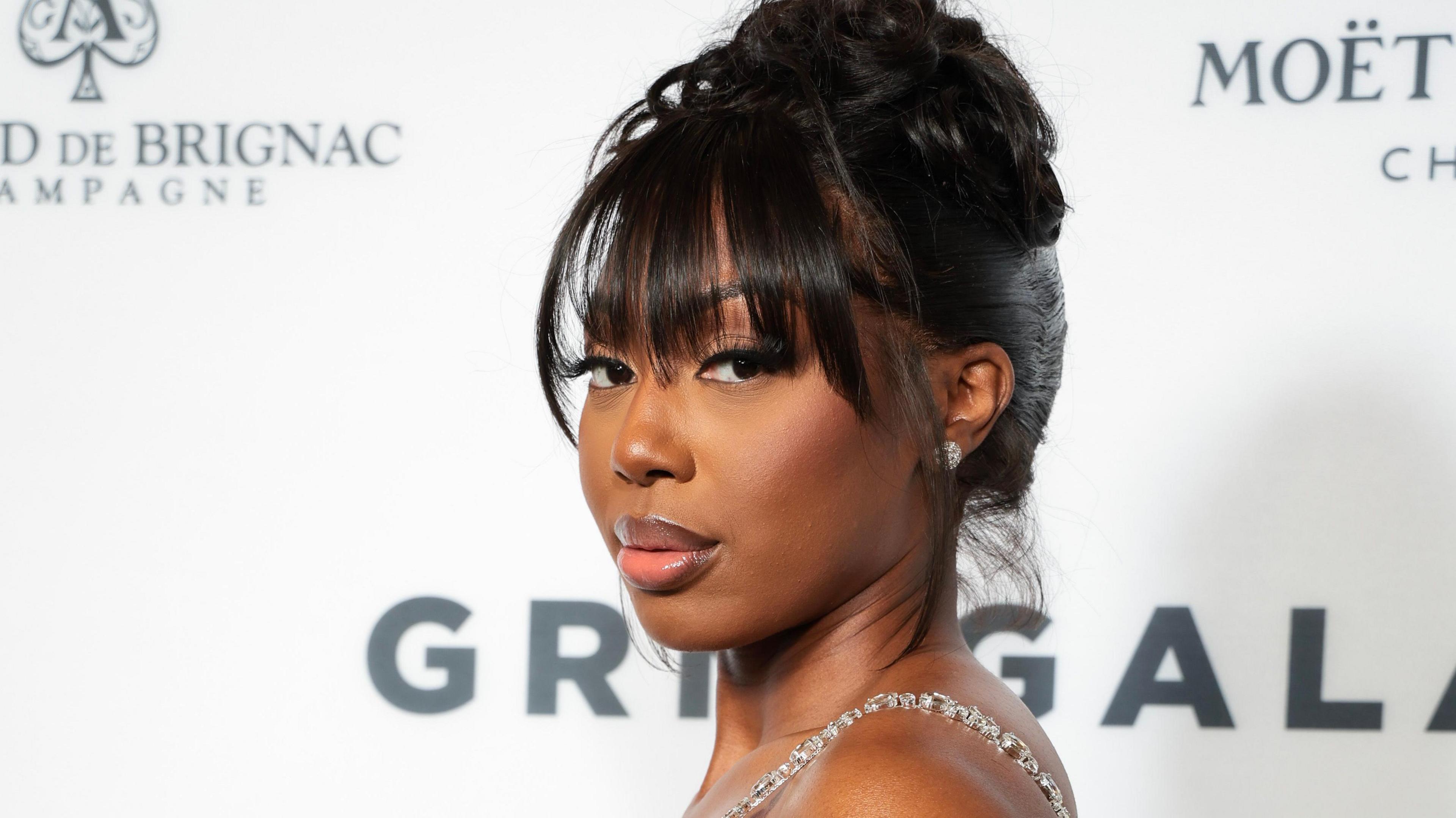 Ms Banks at the GRM Gala in London. Ms Banks wears her hair tied up in a bun on her head with a fringe down past her eyebrows. She looks at the camera over her left shoulder, wearing a jewel embellished bustier dress.