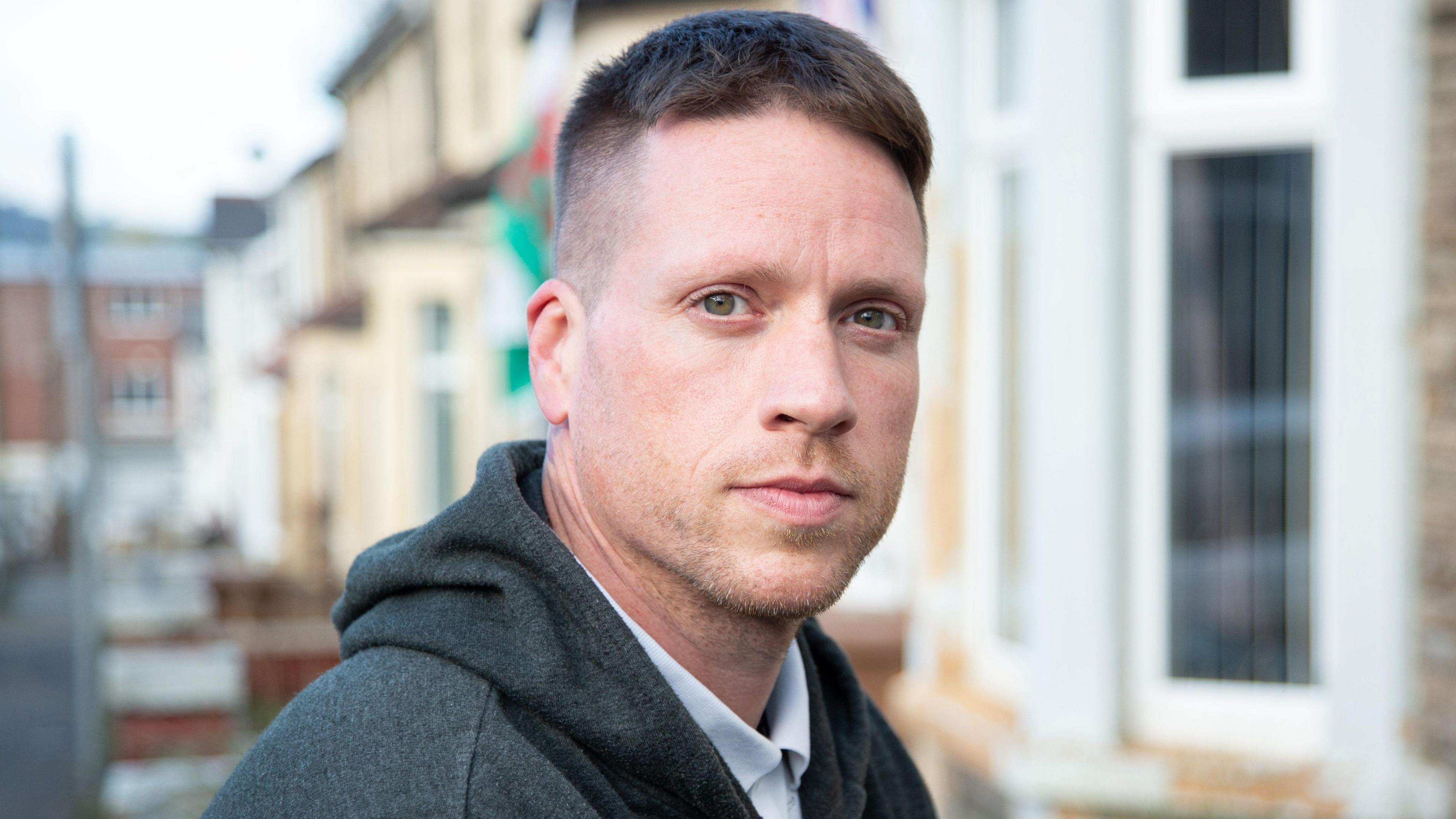 A man with short brown hair and light stubble looks at the camera. Behind him are a row of terraced houses and a Welsh flag.