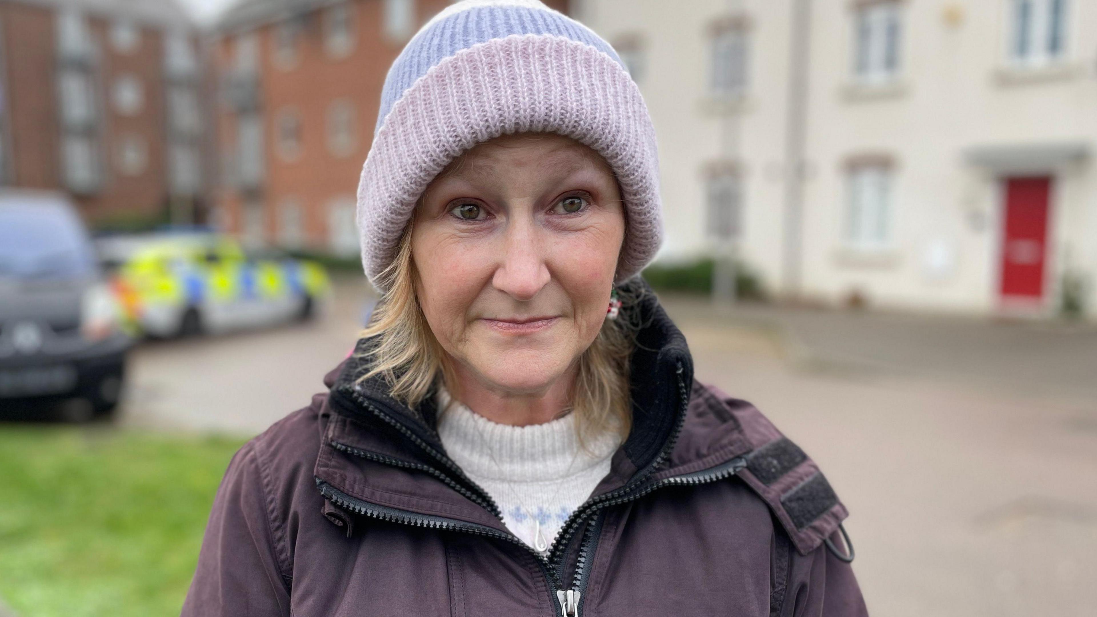 Tracey Howard has tears in her brown eyes and is looking at the camera. She wears a pink beanie hat, white jumper and purple coat. She has shoulder length blonde hair and a silver necklace. Behind her is a police car. 