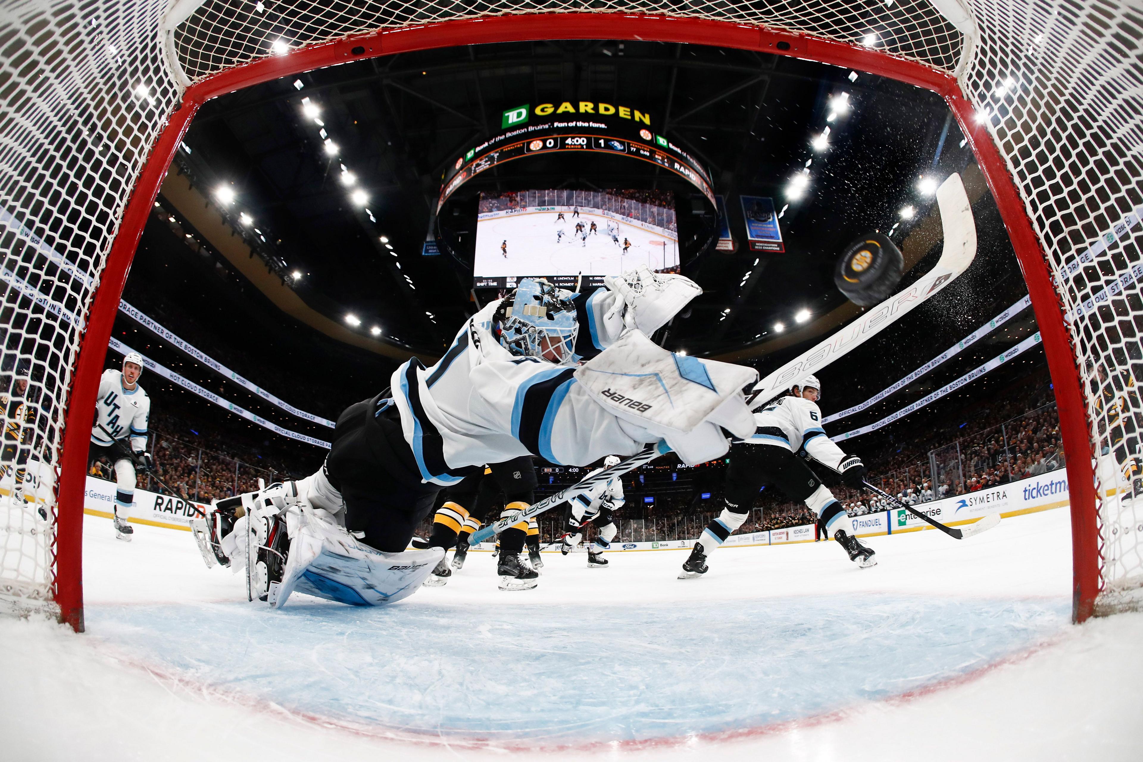 A dramatic low-angle view from inside the goal shows an ice hockey goaltender in white and teal gear fully stretched out, glove extended, attempting to block a puck that flies past the crease. Players scramble around the net as the arena lights shine overhead and the crowd watches intensely from the stands.
