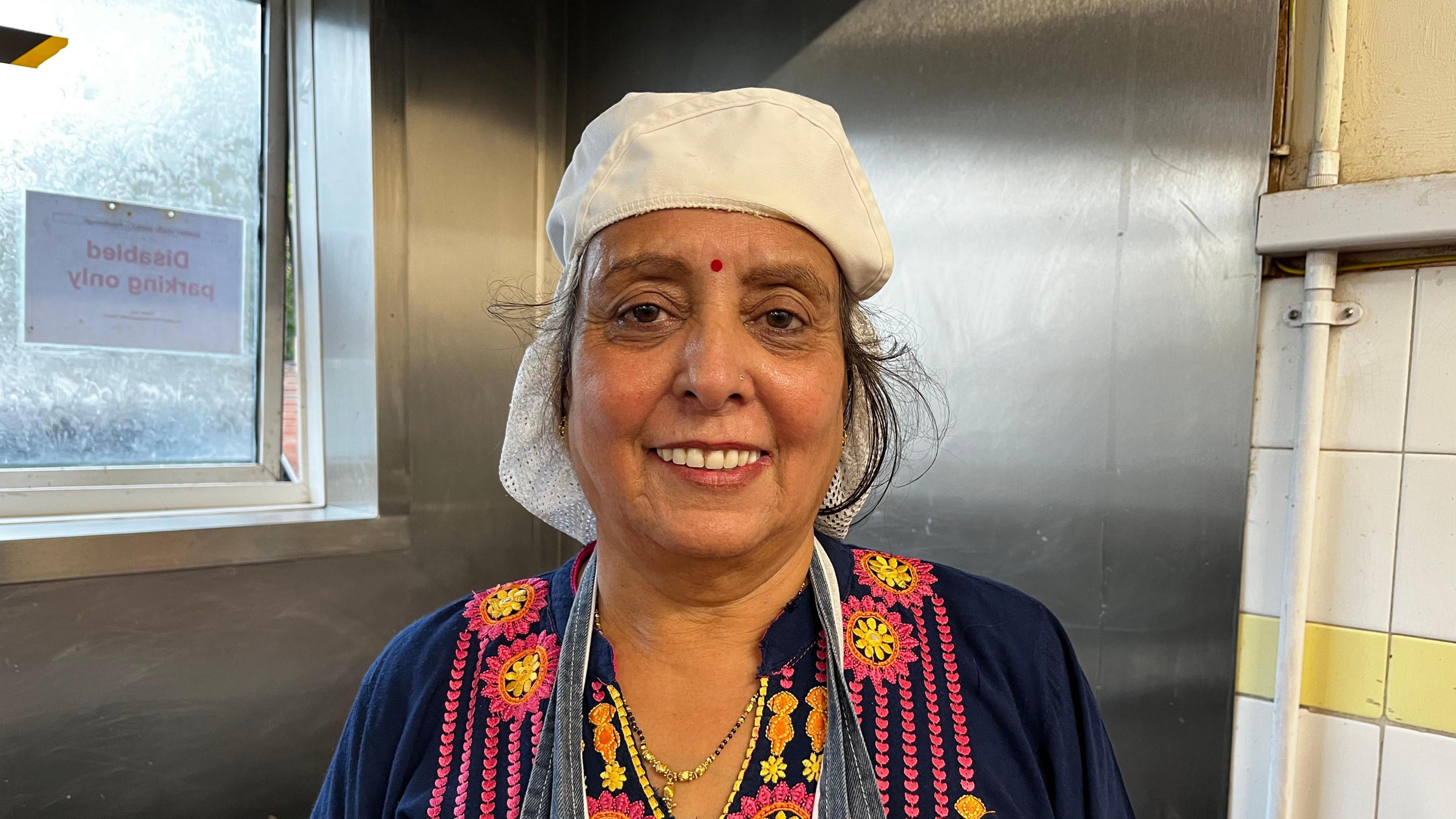 Mina in her kitchen smiling at the camera. She is wearing a white head protection cover, a grey apron and a white and pink kurta.