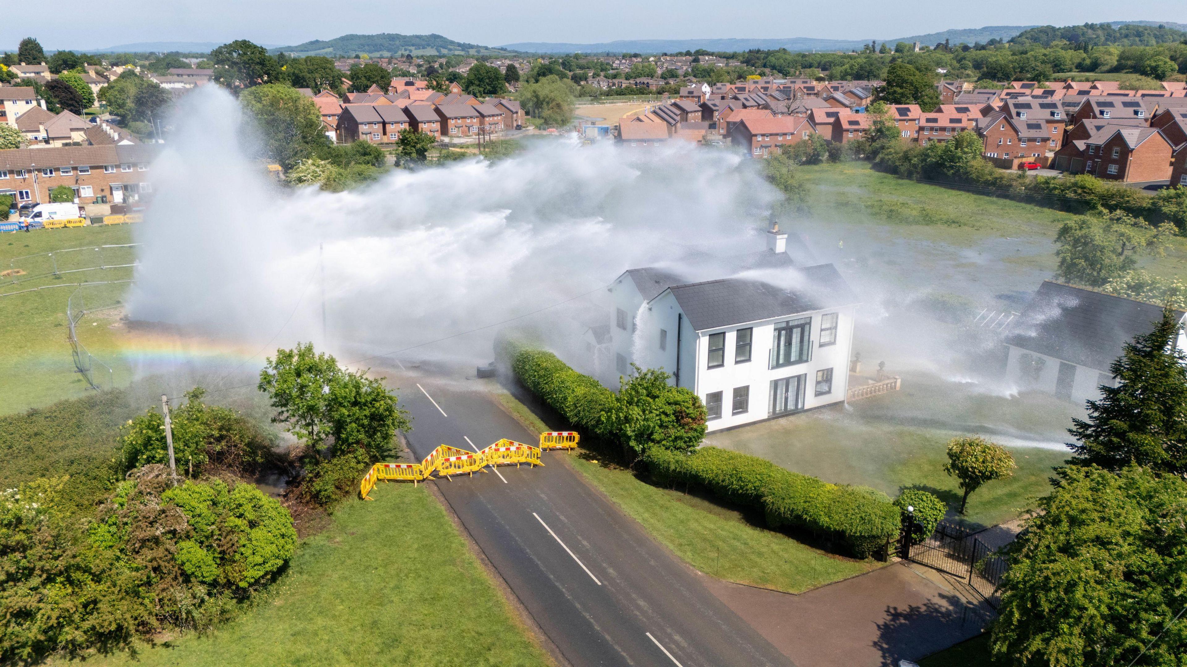 A drone shot of the burst water pipe in Matson, it shows a large torrent of water shooting from a field, across a two lane road and directly onto a large house. The road has been cordoned off with yellow fencing.
