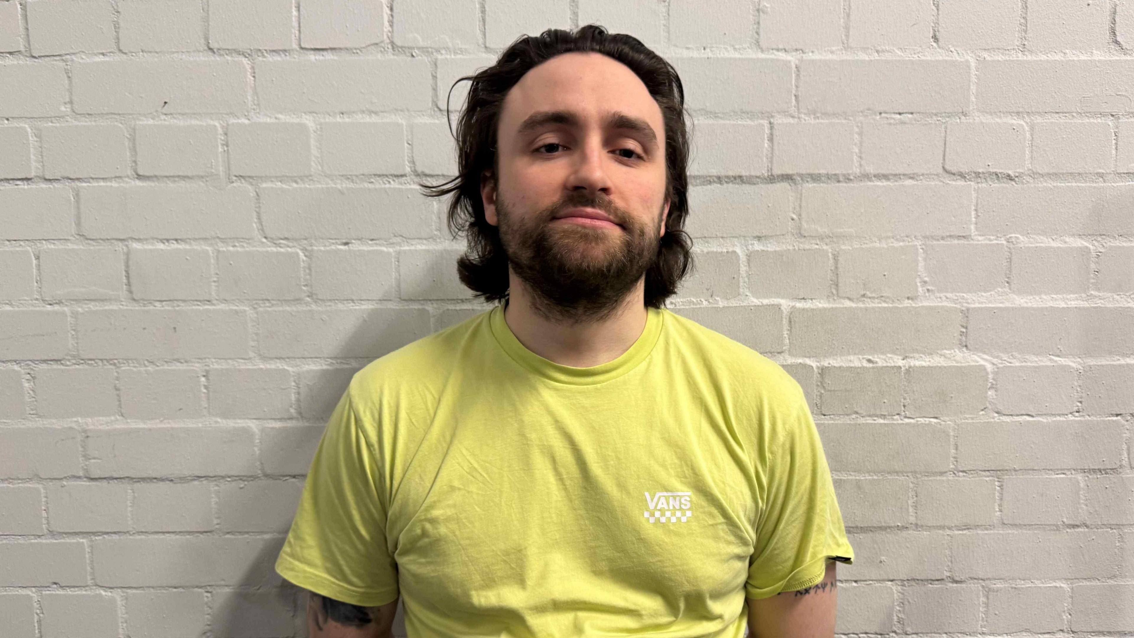 A man with mid length dark hair and a yellow top looking at the camera.