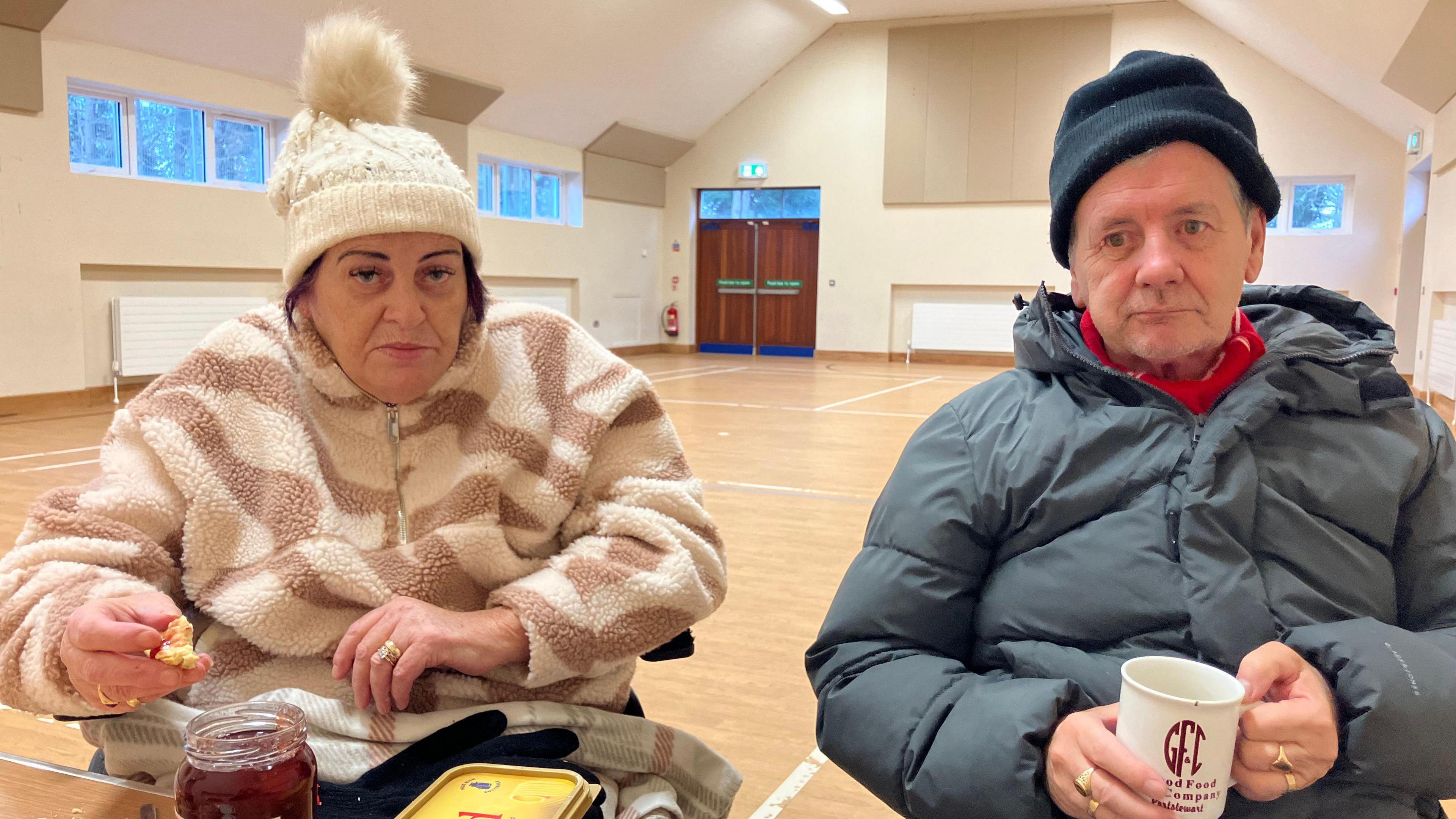 Desmond and Olive Hyndman sit in a community centre. Desmond holds a mug and Olive holds a scone. There is butter and jam in front of them. Desmond wears a grey puffer jacket and grey hat and Olive wears a fleece and cream hat.