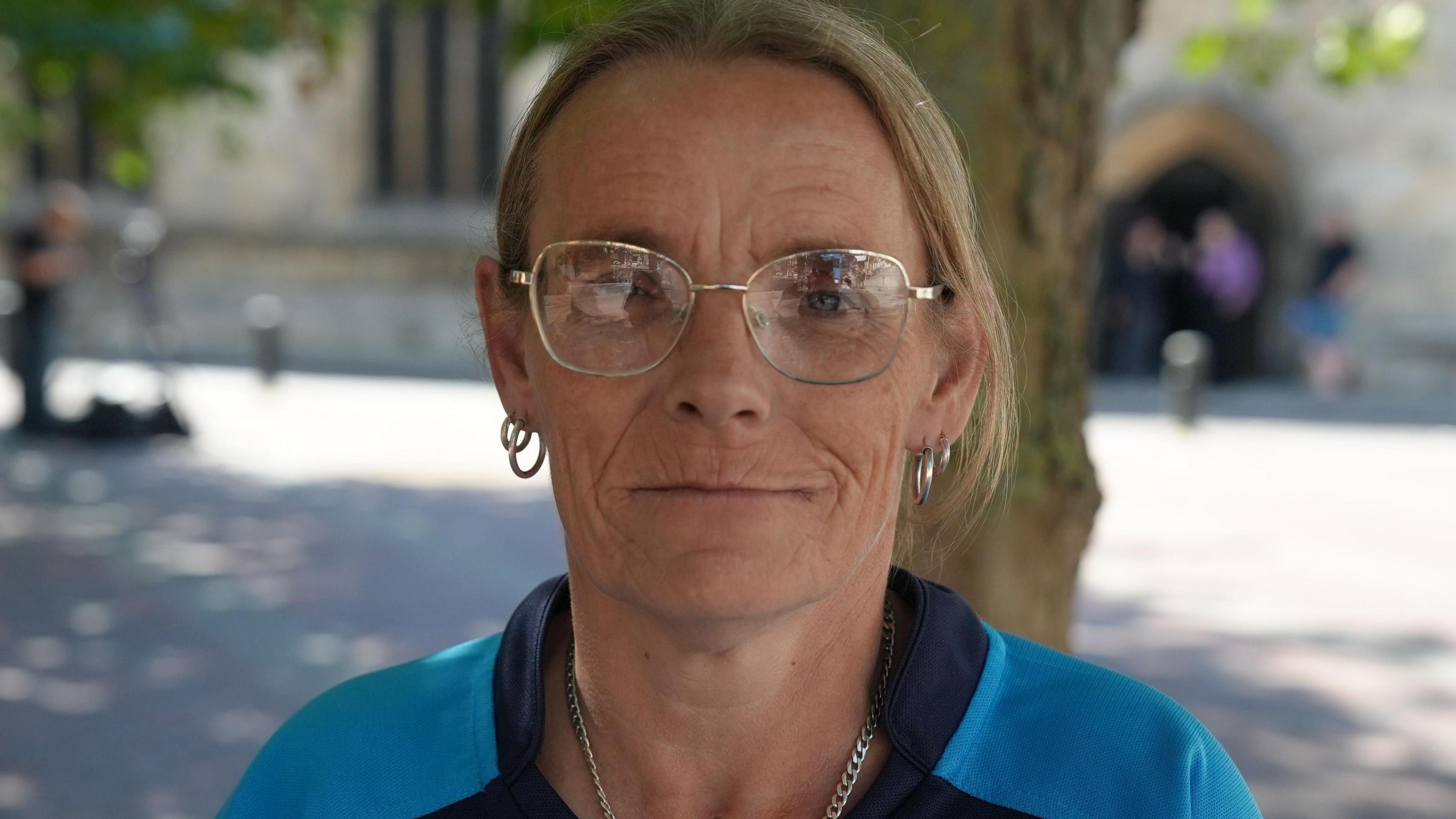 Joanne Moody is wearing large specs on her face, two hoop earrings, a necklace, and a blue sports jersey. The background is softly blurred and includes a tree, courtyard and unidentifiable people in the distance.