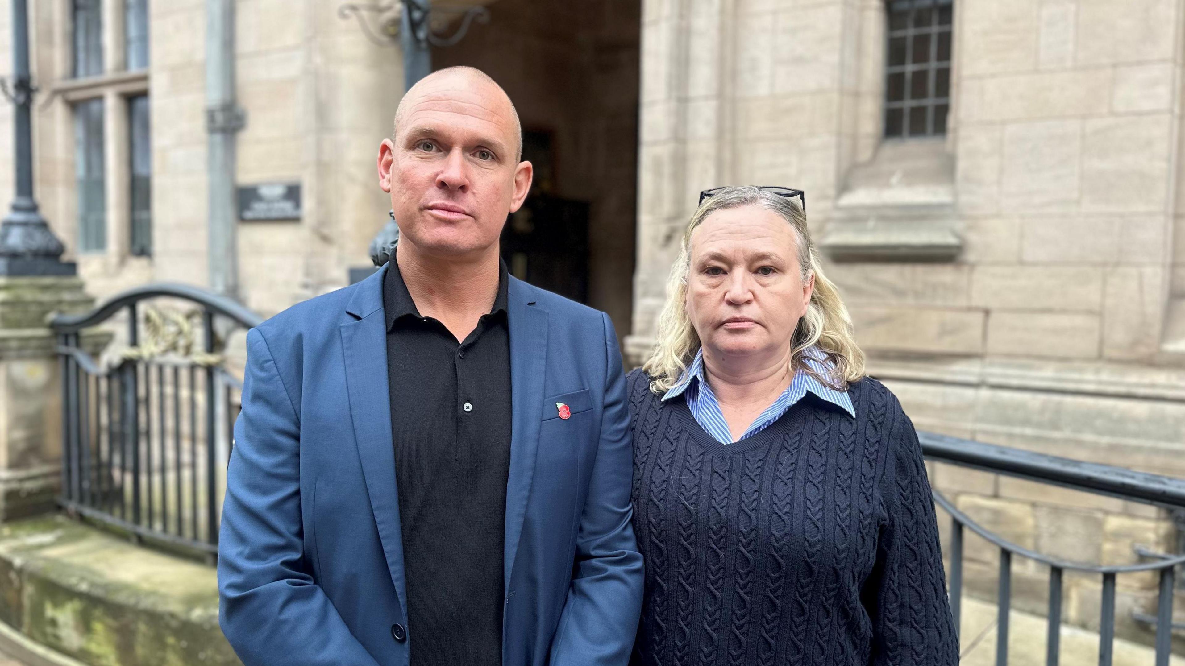 Chris and Natalie Watkiss. Chris is wearing a black polo shirt with a dark blue suit jacket and Natalie is wearing a blue shirt with navy blue jumper. They are standing outside a stone building which has black railing alongside it.