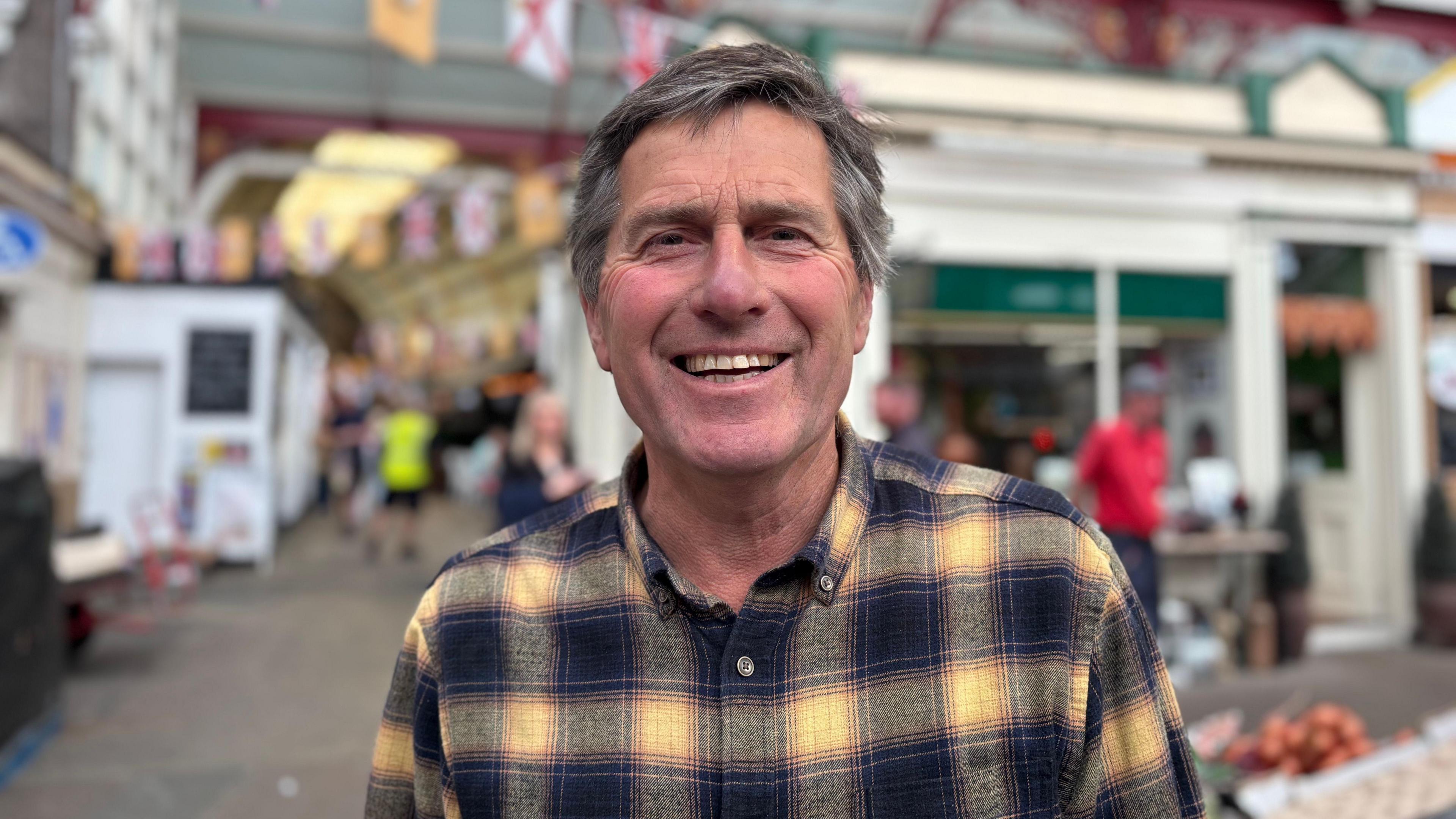 A middle-aged white man smiling at the camera. He has dark grey hair and is wearing a yellow and black checked shirt.