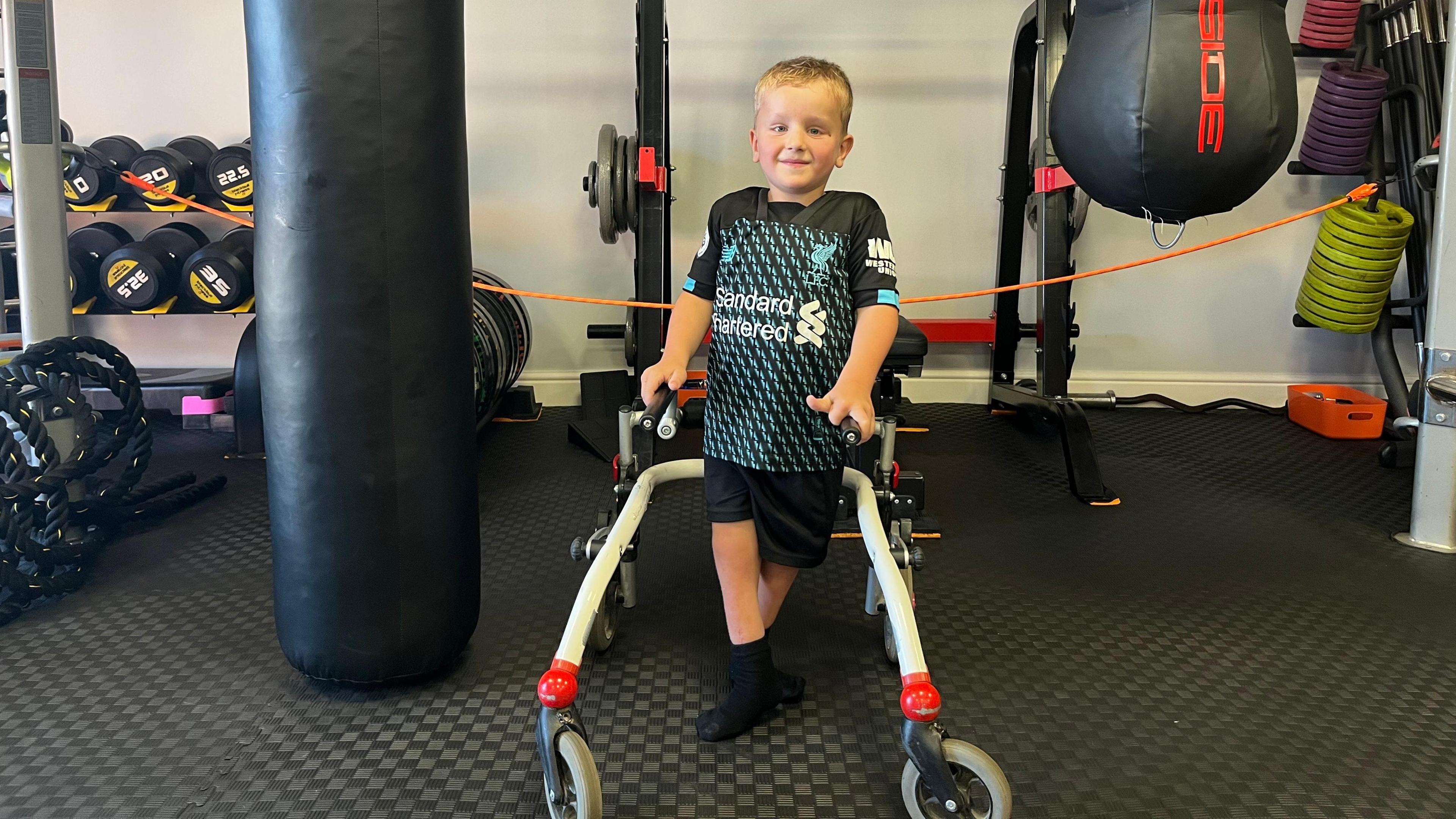 Enzo is holding a walker to support himself and his in between two punchbags ib the gym