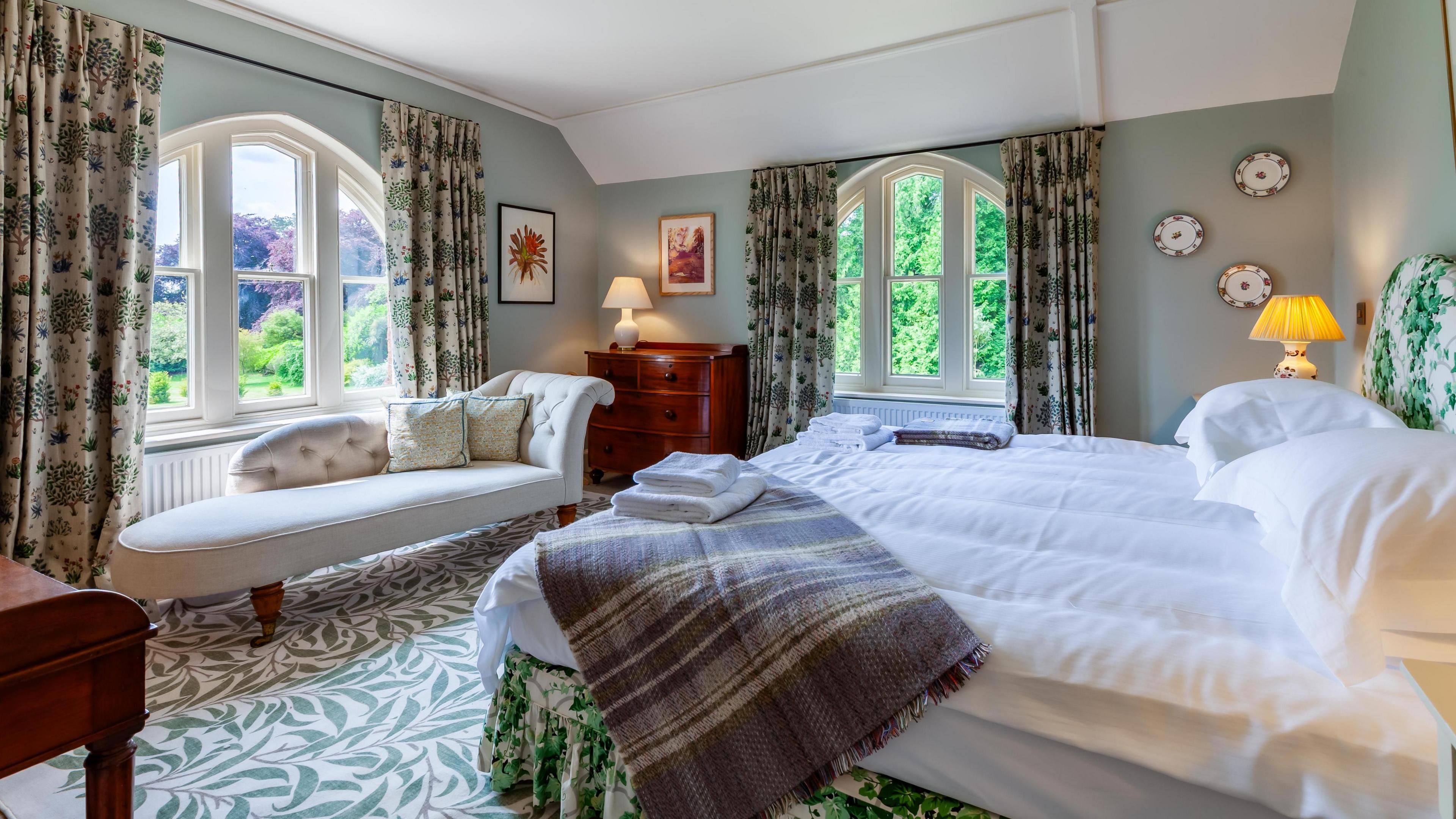 Master bedroom which has a carpet patterned with green leaves, a large double bed, a chaise long and big windows with views onto the garden.