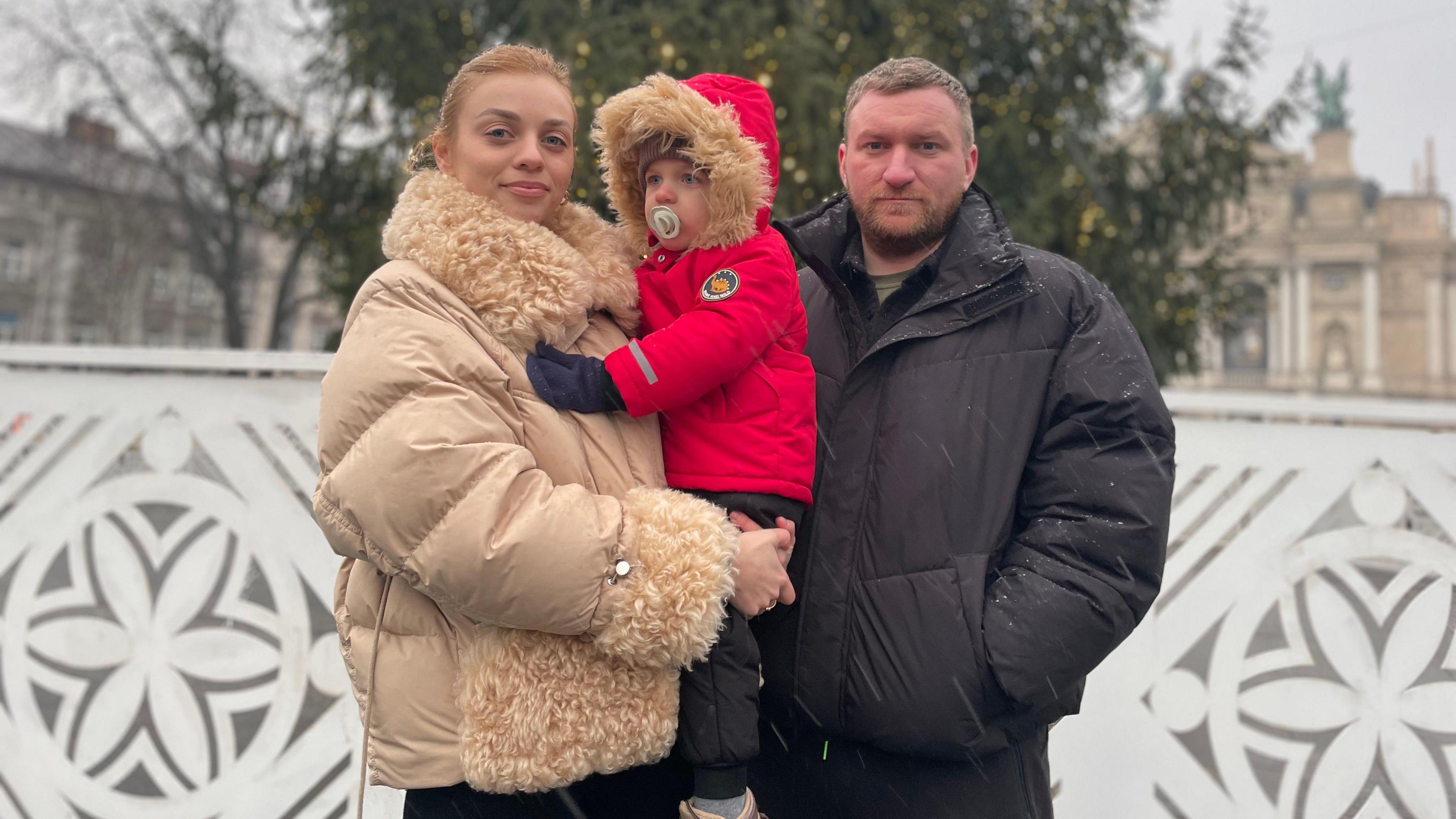 A woman holds a young child in a red jacket and a man stands next to her