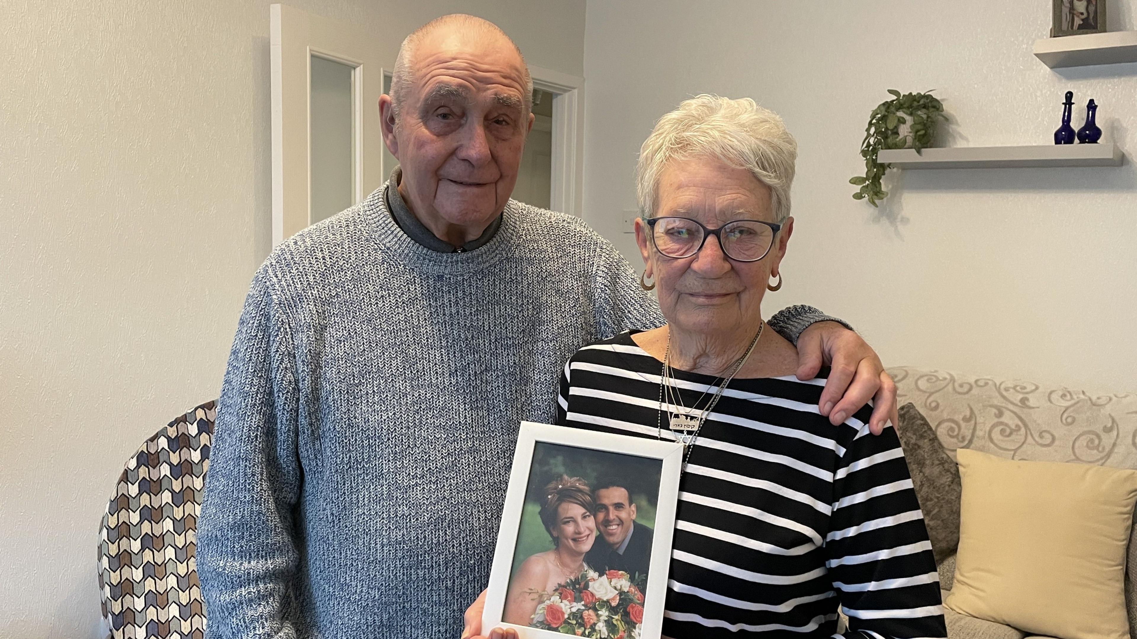 Gill and Pete Brisley looking at the camera holding a photo of Eli and Lianne Sharabi on their wedding day