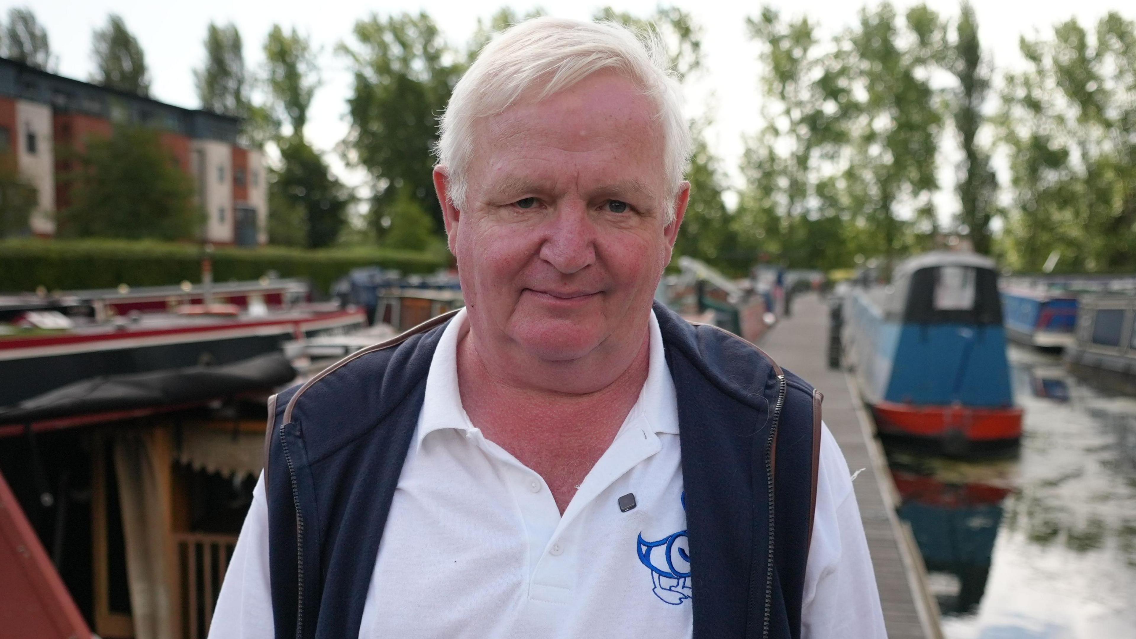 A man standing on a dock with moored boats in the background. He is wearing a white polo shirt with a logo and a dark vest.