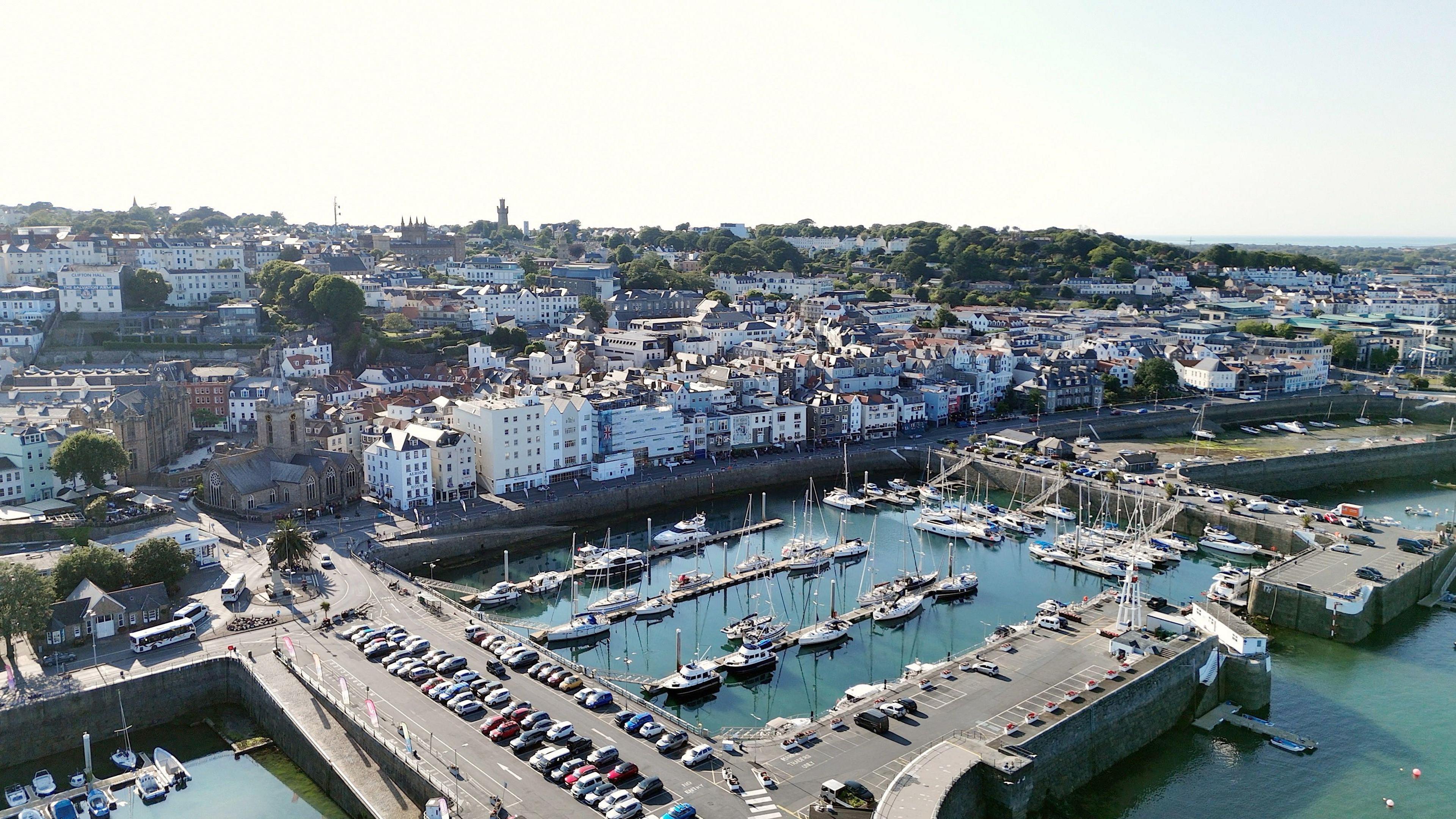 An aerial photo of the harbour and St Peter Port on a bright day. There are many yacht and tenders moored up and the harbour car park looks full. 