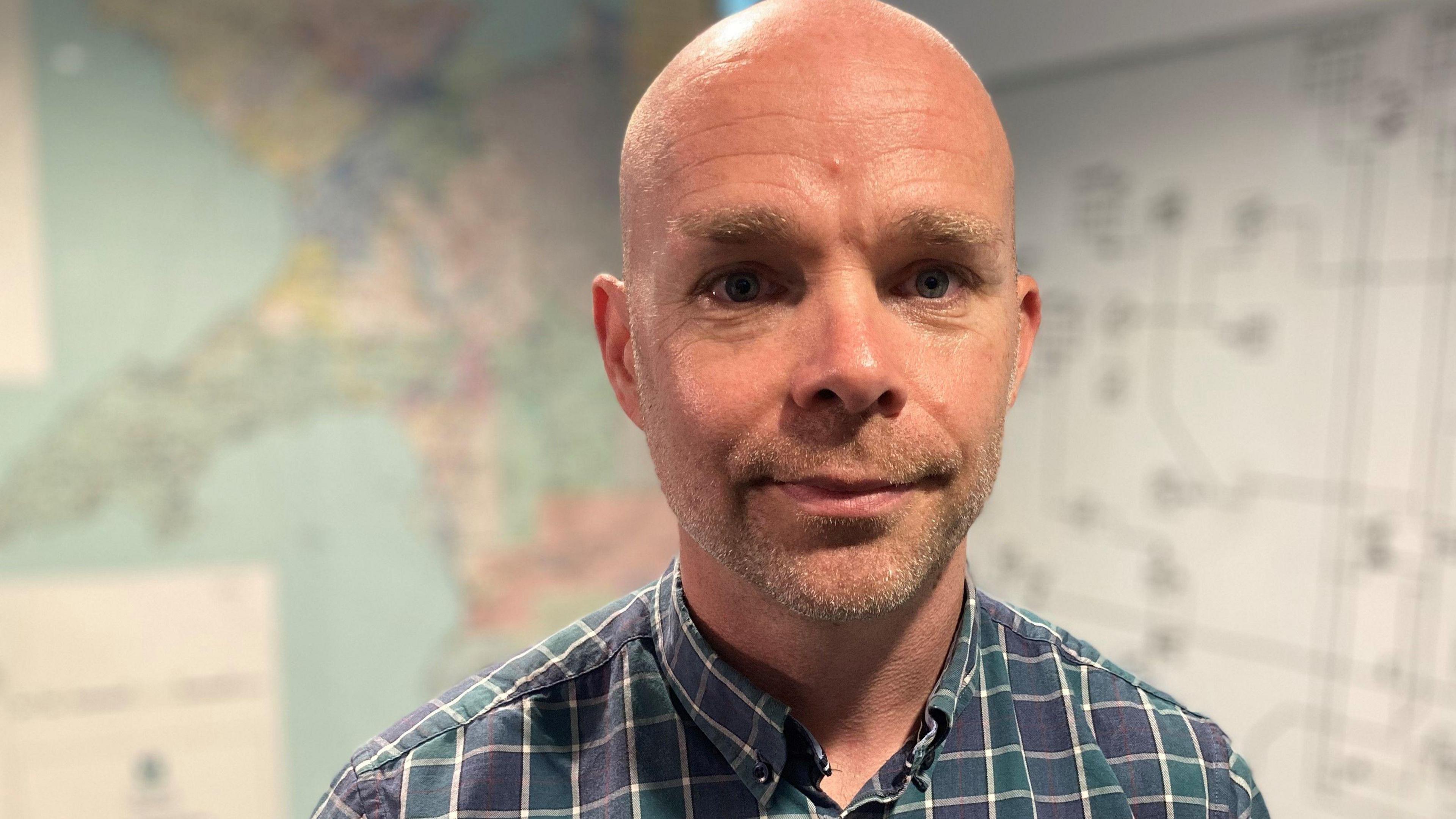 Kit Wilson, director of operations at Welsh Water, stands in front of a map of Wales and a diagram of the water network, wearing a checked blue and green shirt.