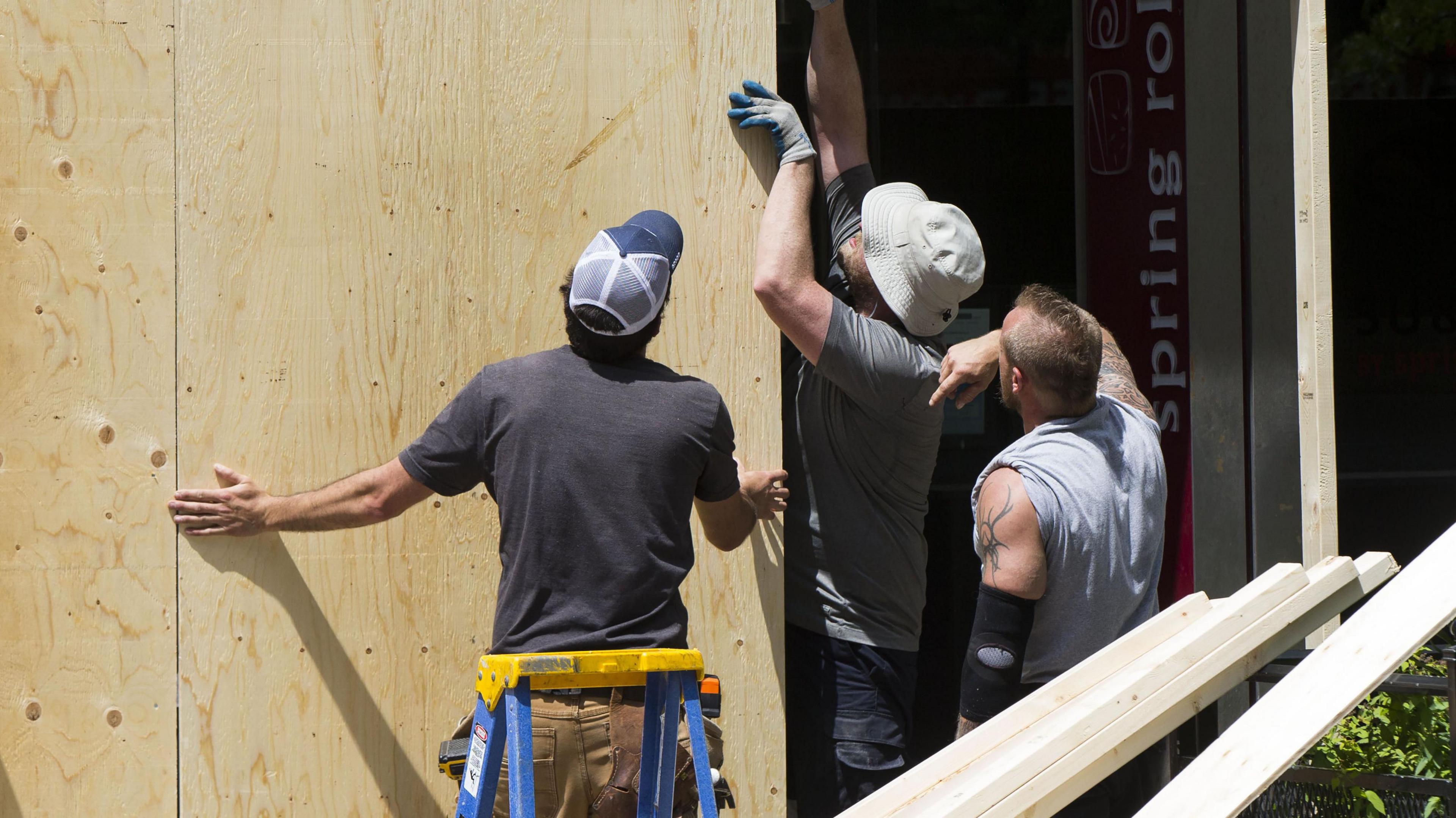 Three builders are looking at a large wooden panel in a shop window on a street. Two of them have their hands on it and the other has his arm on one of men's shoulders. The men wear T-shirts and hats and there are long lengths of wood in the foreground.