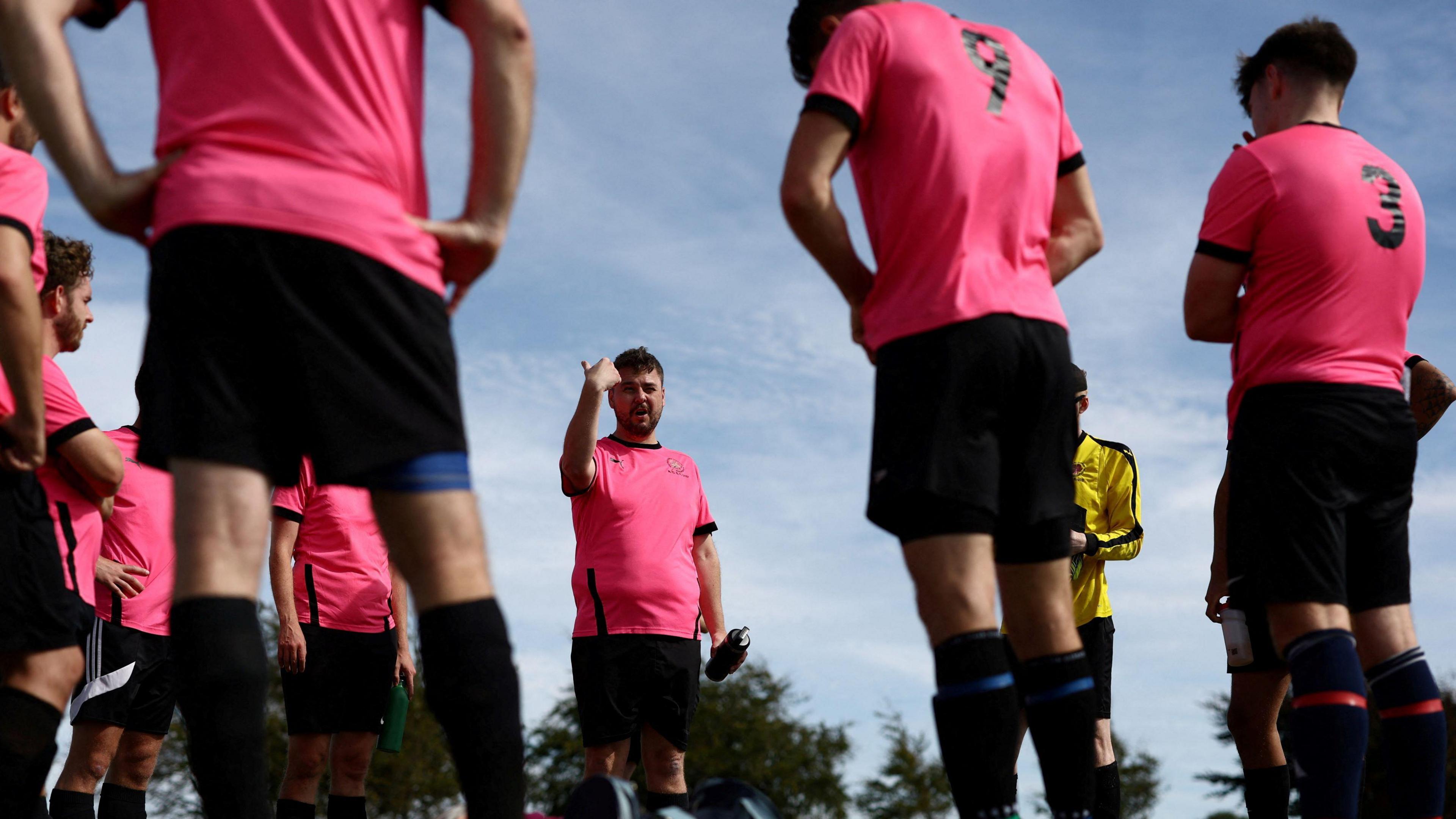 A group of male footballers wearing pink tops and black shorts stand in a circle as one player, who has dark hair and a beard, gives them instructions before a Downs League game in Bristol.