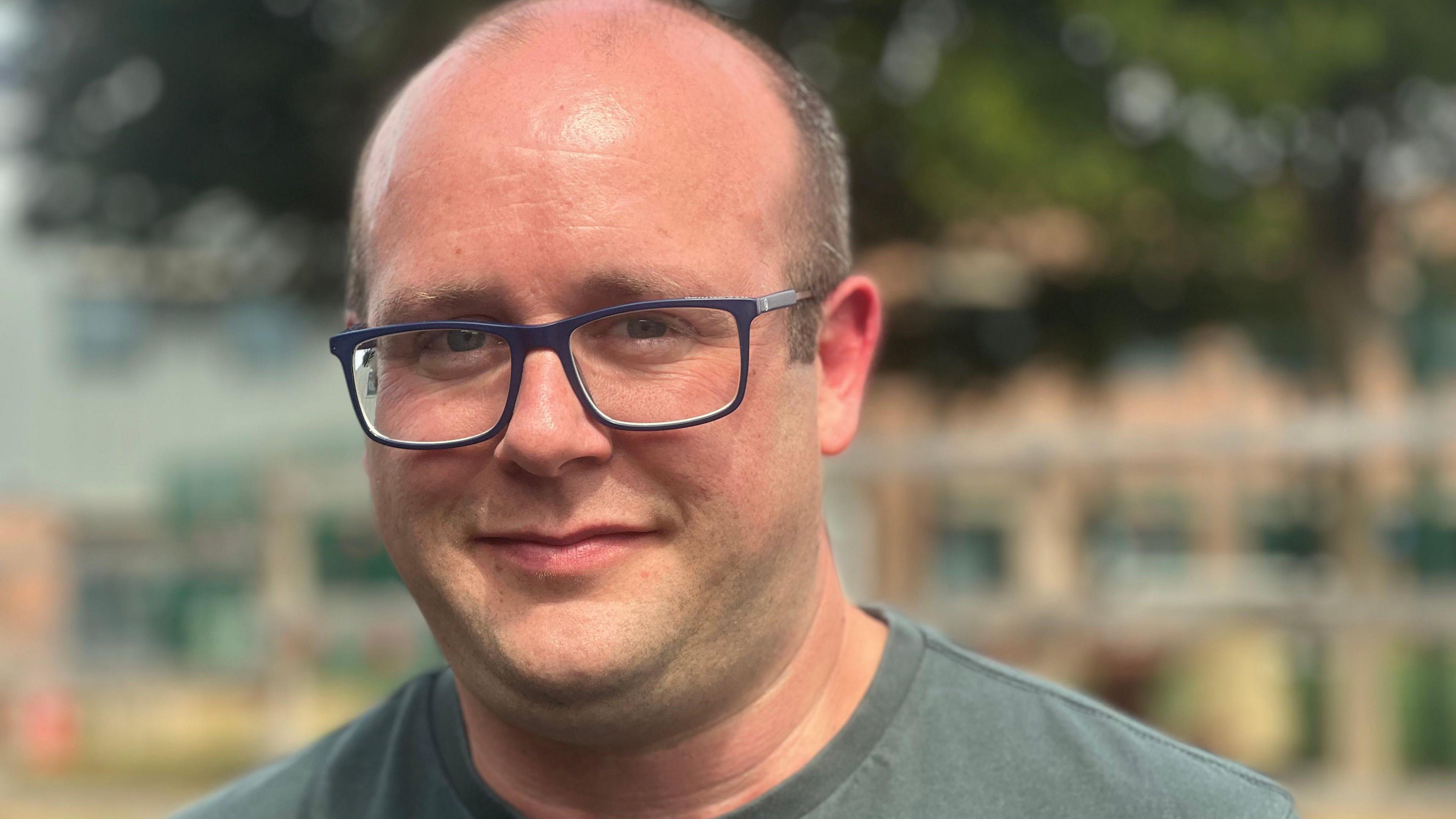Ed smiles at the camera with his mouth closed. He wears square blue-framed glasses and a dark green t-shirt.