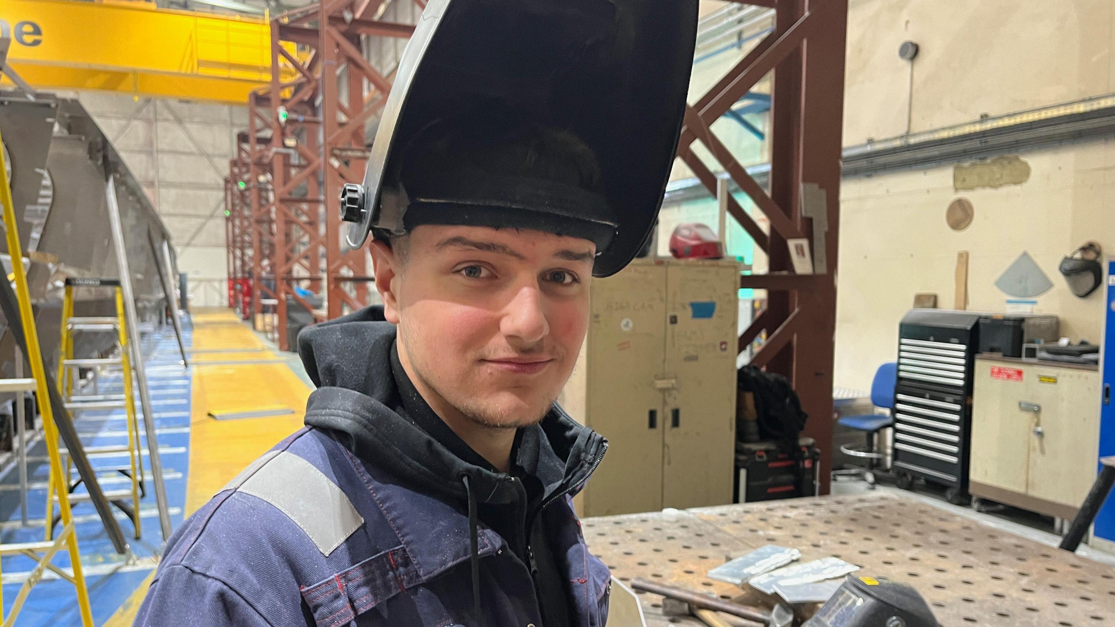 Zak Linington is wearing blue overalls and has a welding mask cocked above his face - behind him is a workshop.