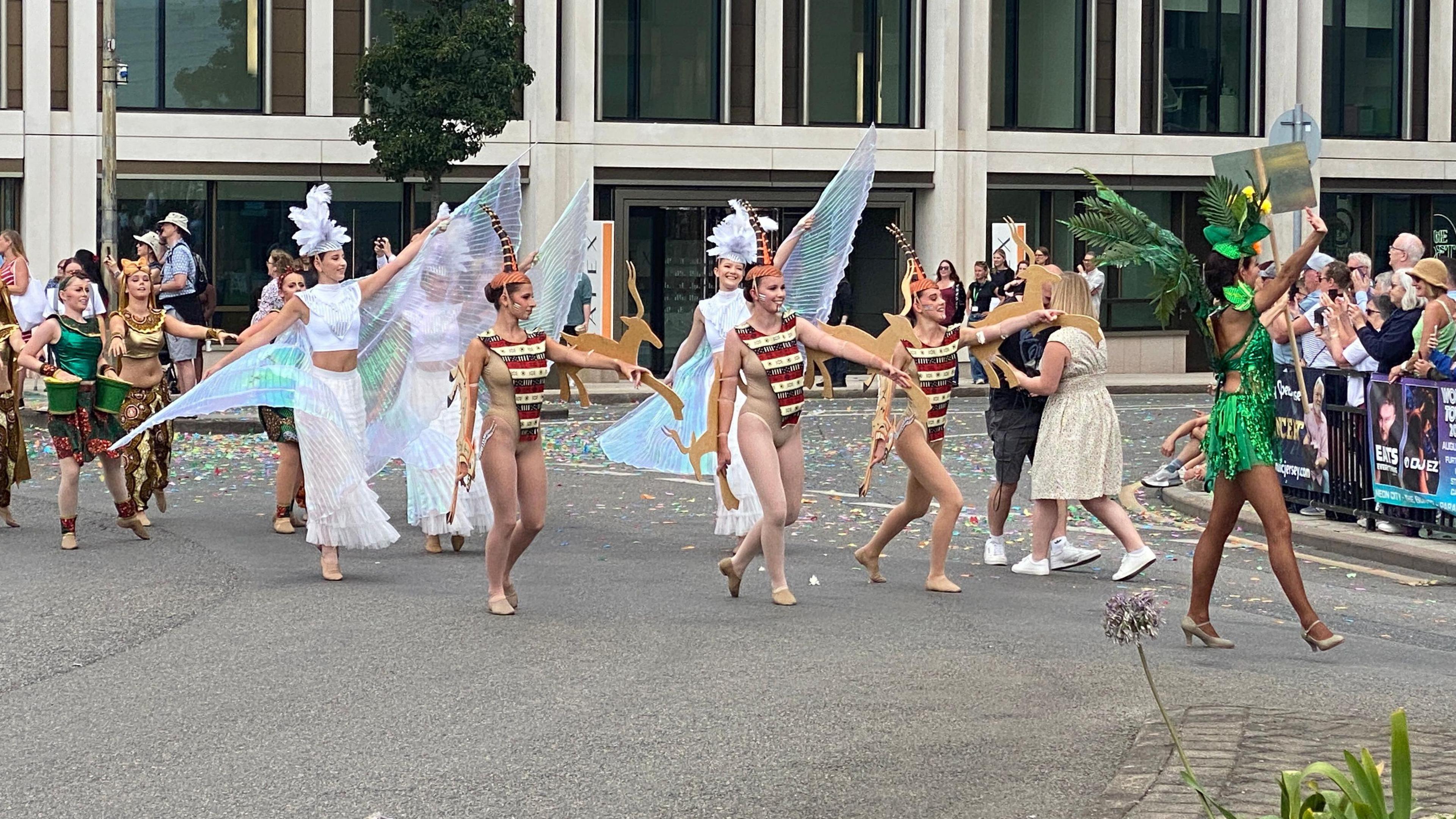 Eight girls are dressed up and are walking in formation. At the the head of the procession is a woman dressed in a short dress of green sequins and palm leaves. Behind her are three girls wearing flesh-coloured bodies with patterned short sleeved ponchos over them. Behind them are three girls dressed in long floaty white skirts who have ethereal wings stretching from their arms. Behind them are three girls wearing shiny brocade tops and patterned skirts.