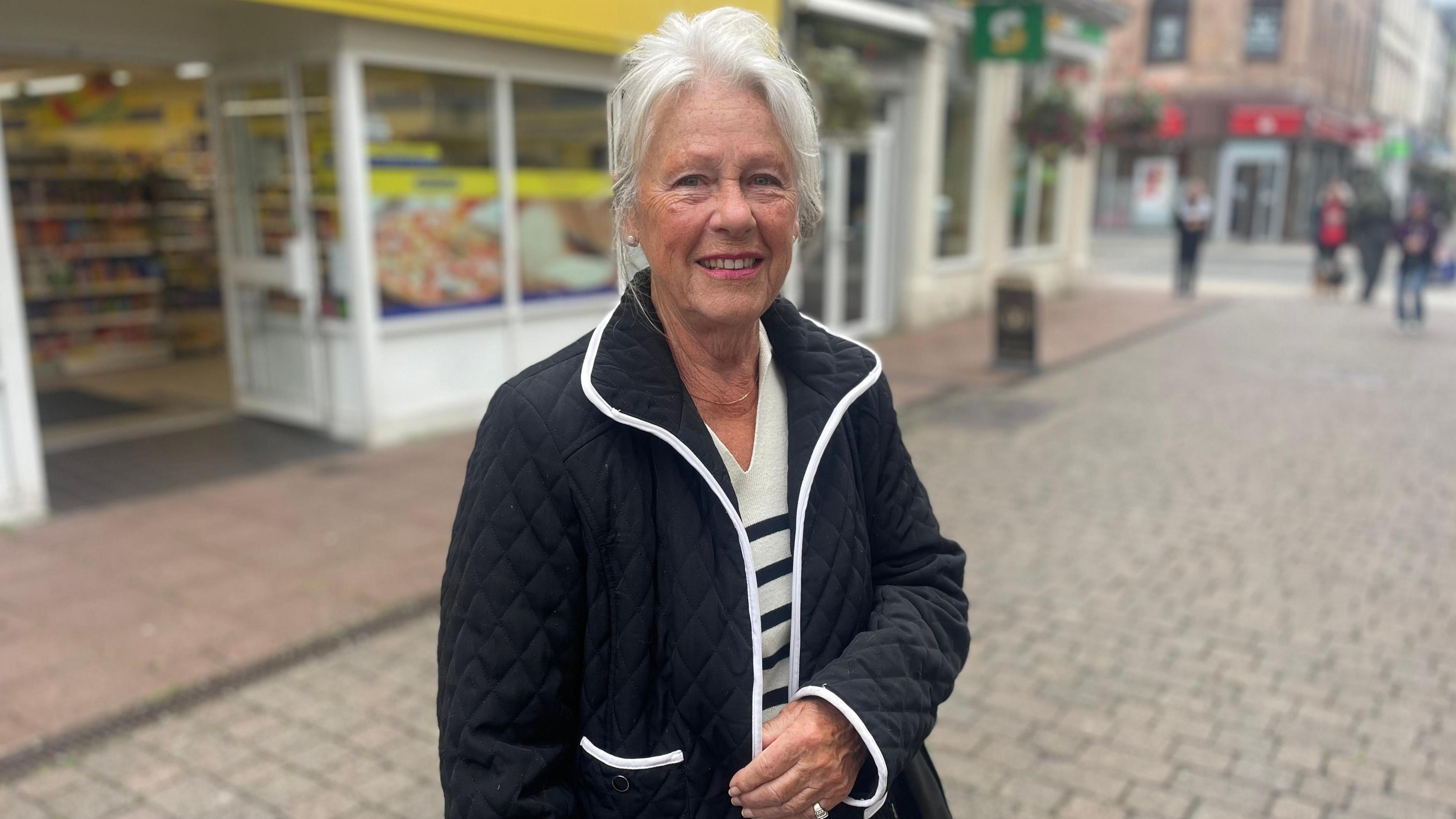 Linda Cuthbert wearing a black and white outfit smiling at the camera on a street in Whitehaven with blurred background.