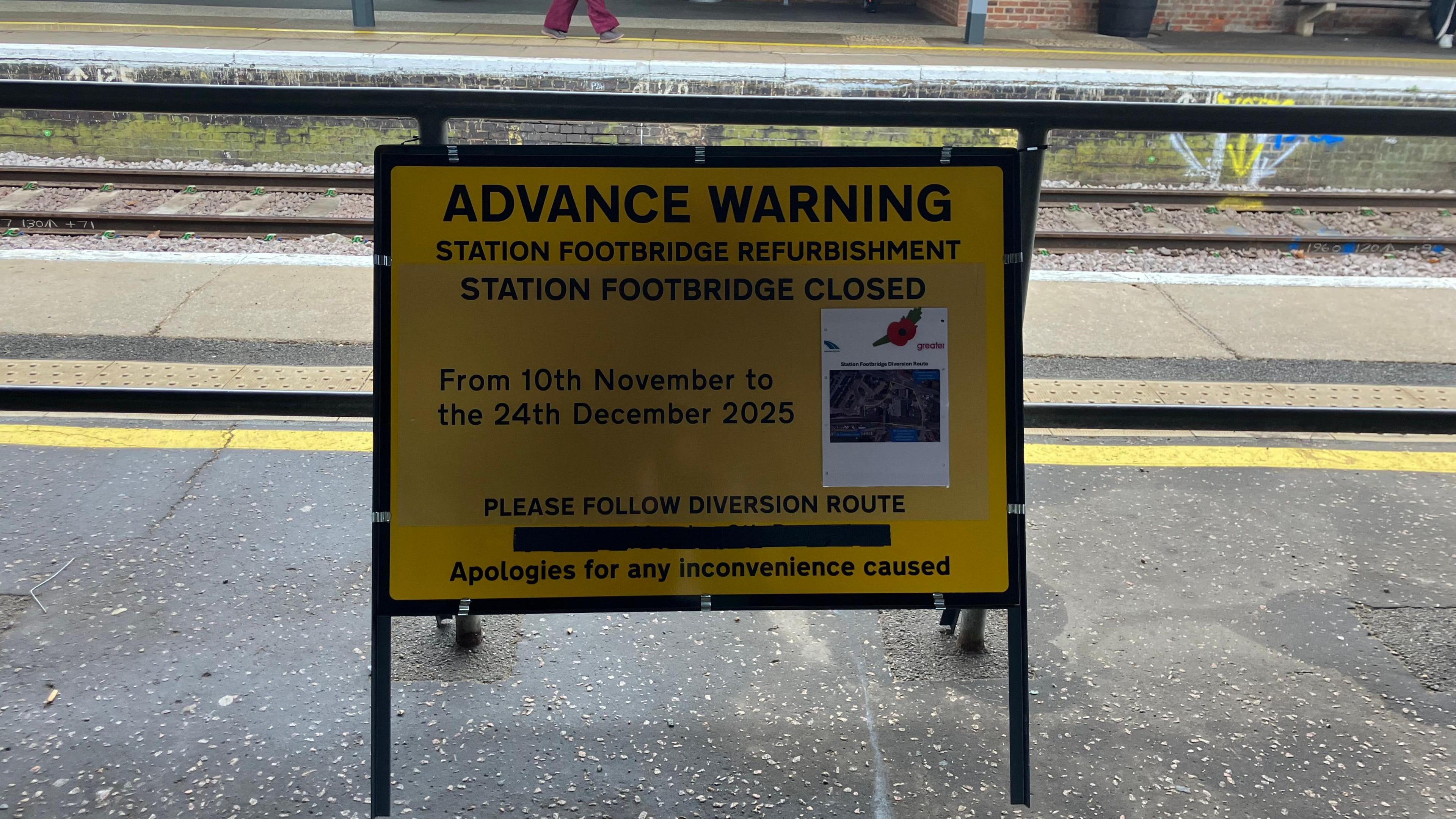 A yellow sign sits on a platform. It gives an advanced warning of footbridge refurbishment works and the dates they will take place. 