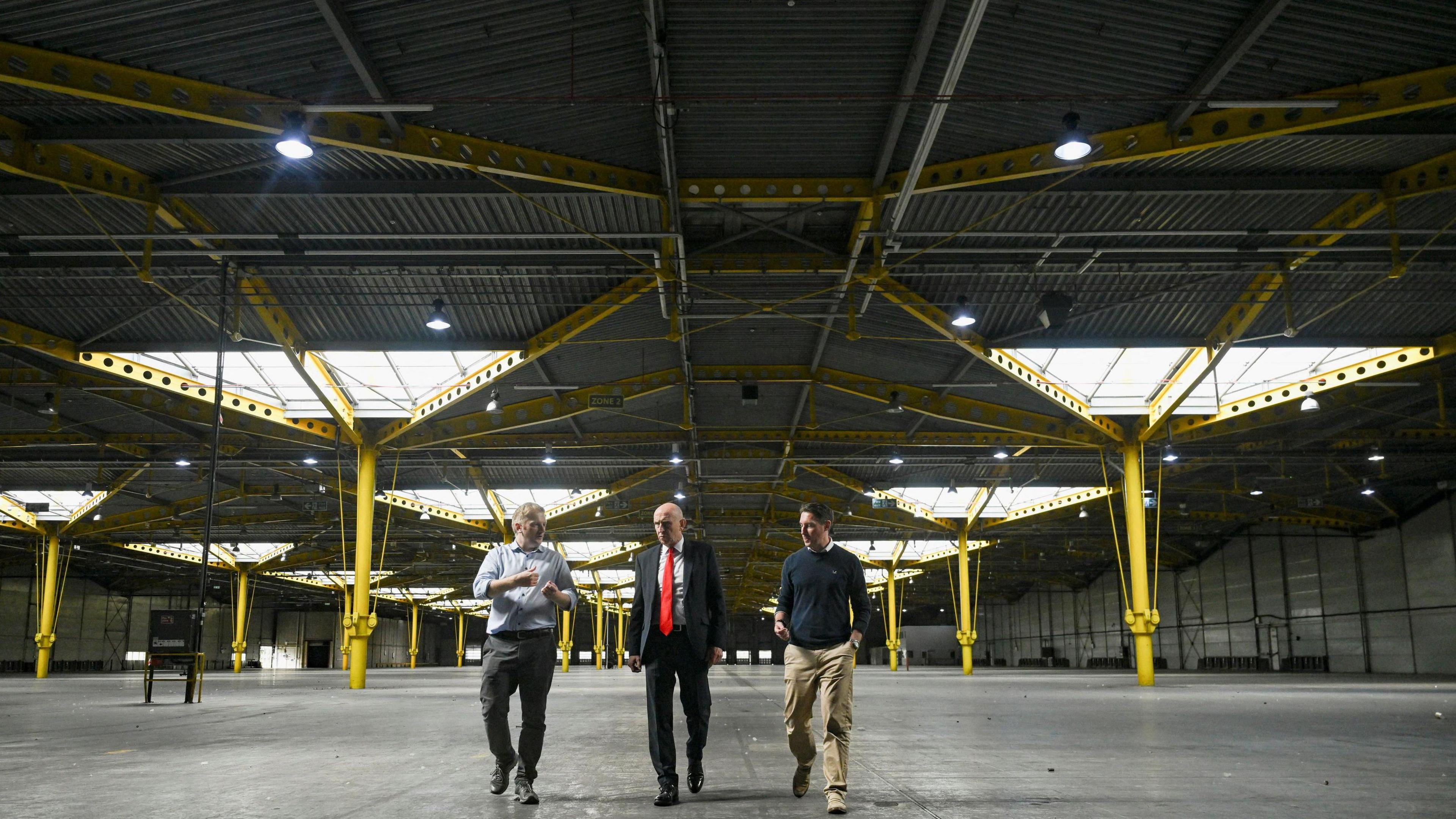 Three men walking through a large empty building space, which looks like the inside of a warehouse and has large yellow columns. 