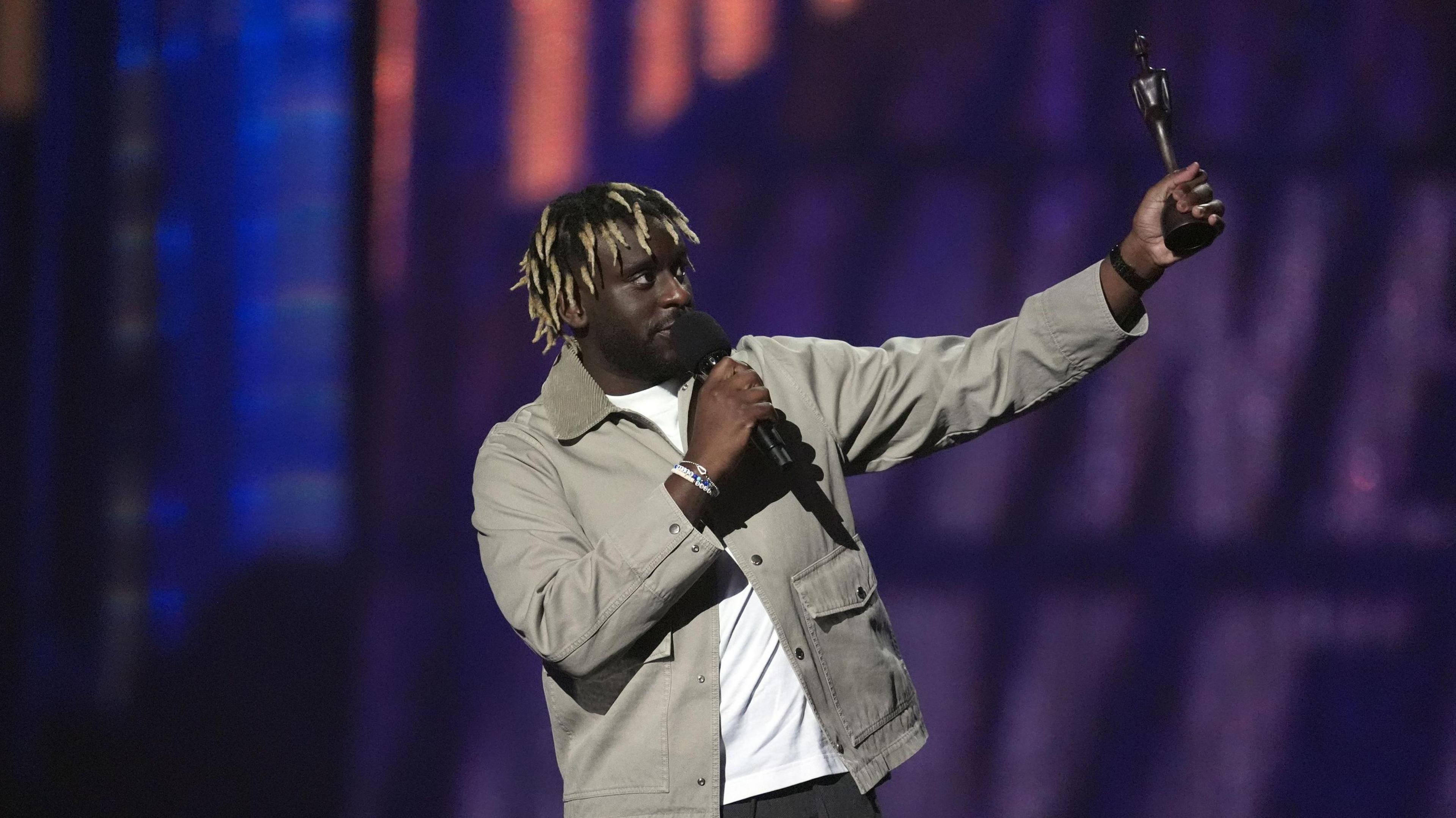 Myles Smith standing in the middle of the picture holding his Brit Award, and speaking into a microphone. The award is the shape of a person, and is made of wood. He has brown and blonde hair, a white tshirt, and beige jacket on. 