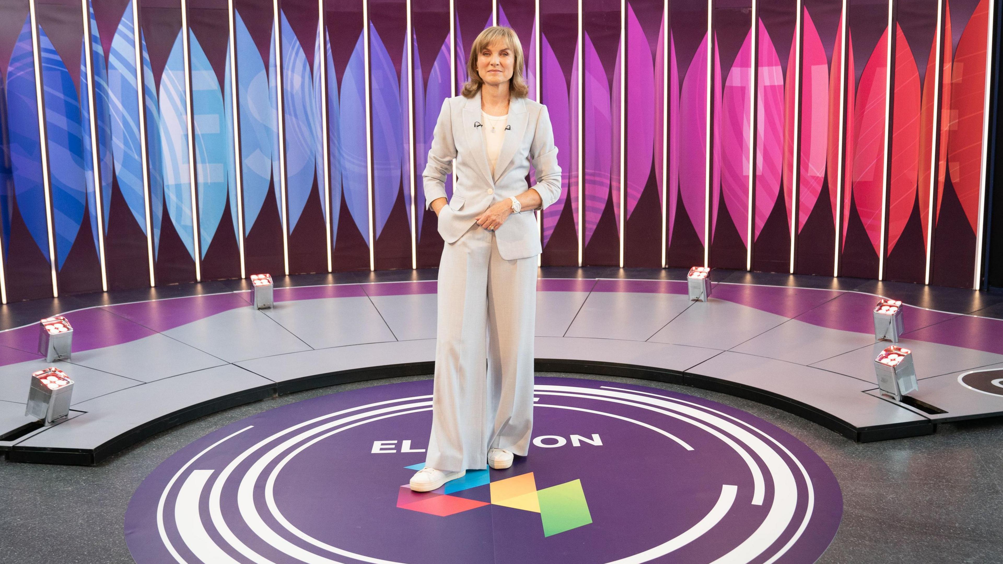 A woman in a grey suit and white trainers stands on a purple circle in the middle of a television studio 