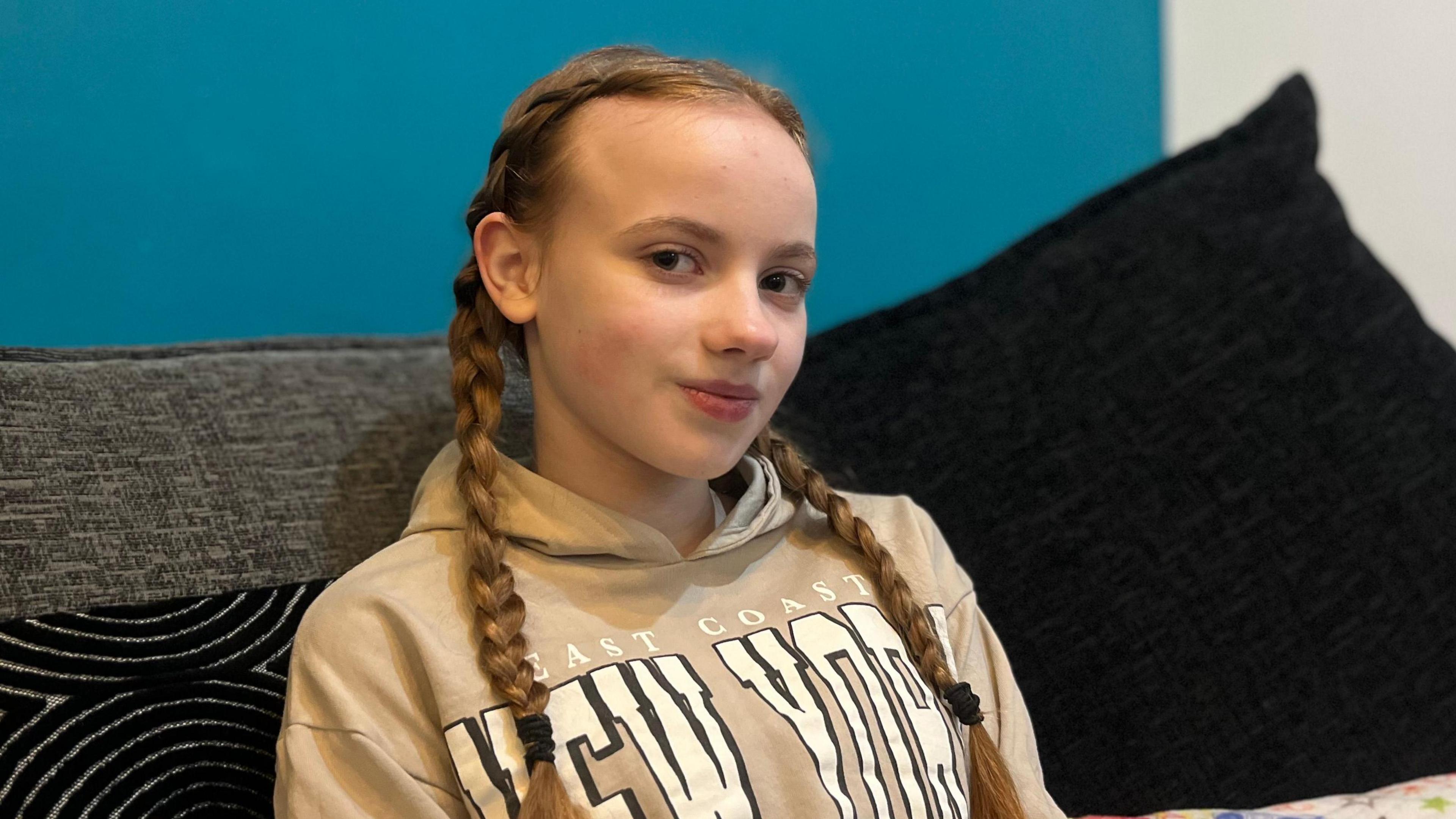 Penny-May is sitting on a dark sofa. She has brown hair in plaits and is wearing a beige New York hoodie.