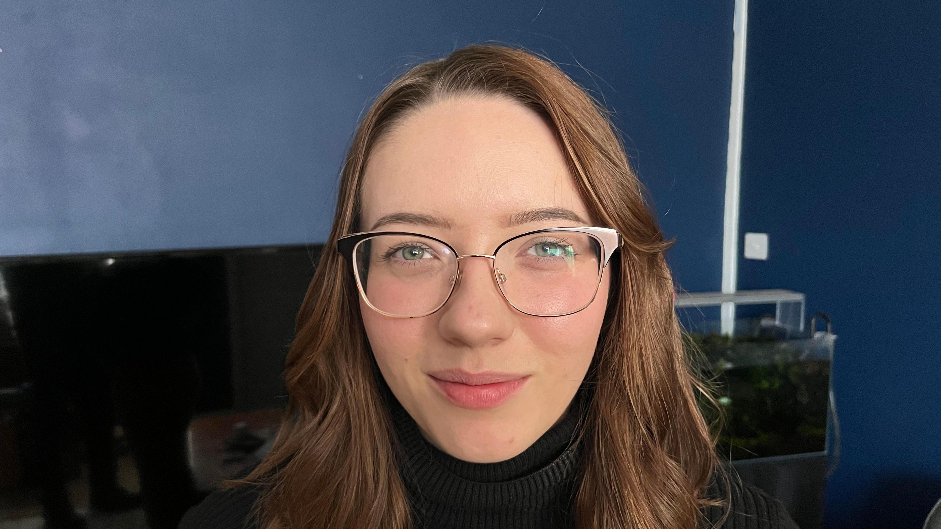 Nicola Ibbotson has straight, brown, oarted, shoulder-length hair. She is wearing glasses with a brown frame and behind her is dark blue living room wall.