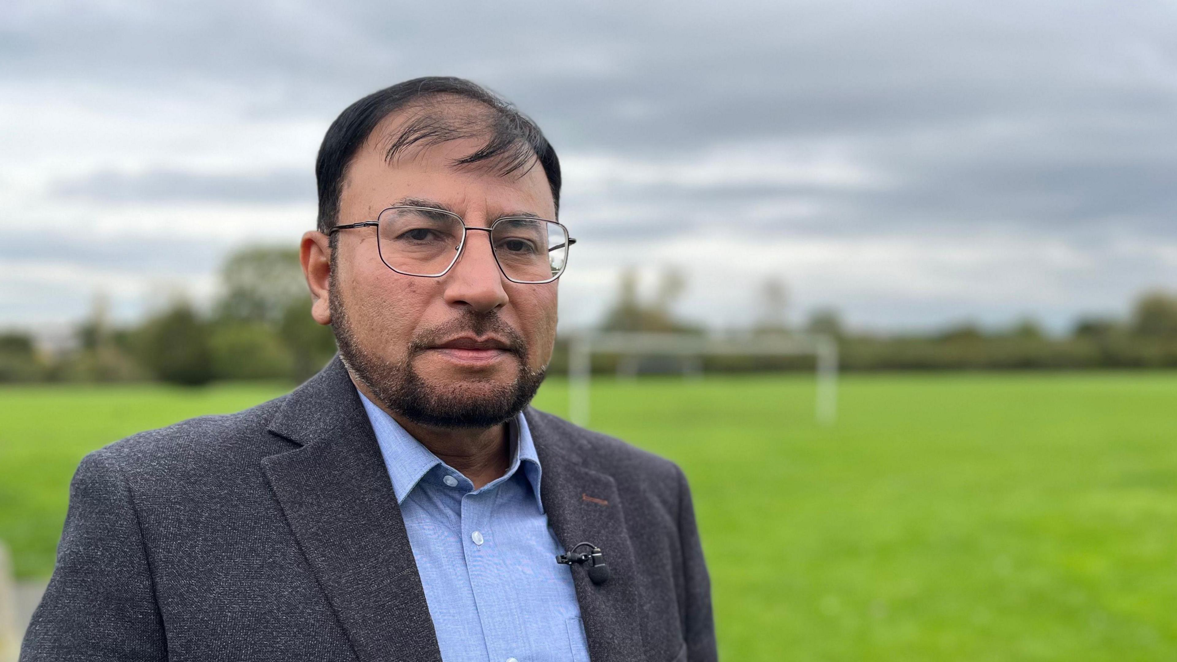 Dr Shakoor looks solemn as he looks directly at the camera. He is standing in a field, wearing a jacket and blue shirt with a microphone attached to his left lapel. He is wearing glasses.