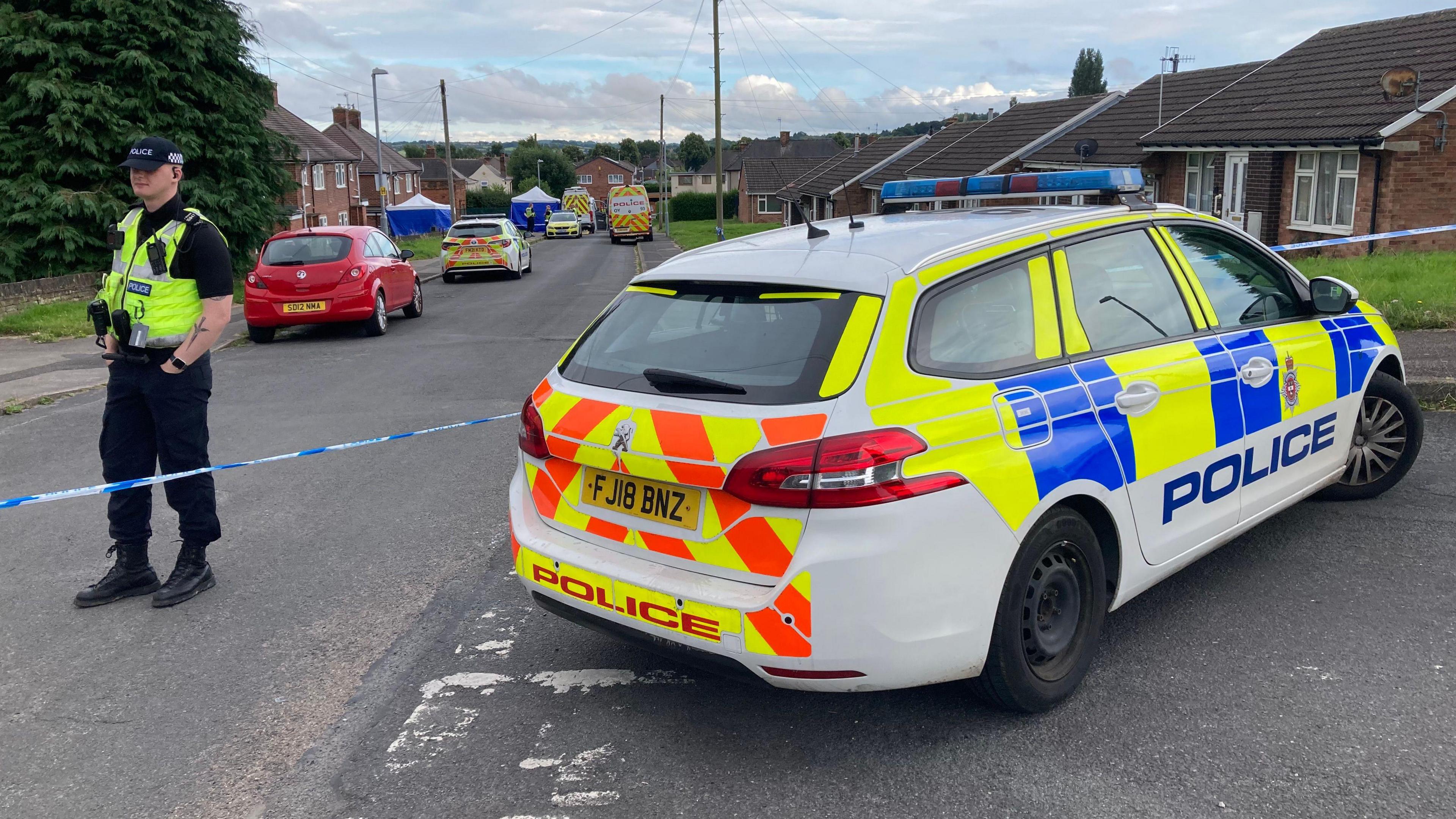 A police officer stands by a cordon and police car on a street with a car and several police vehicles within the cordon