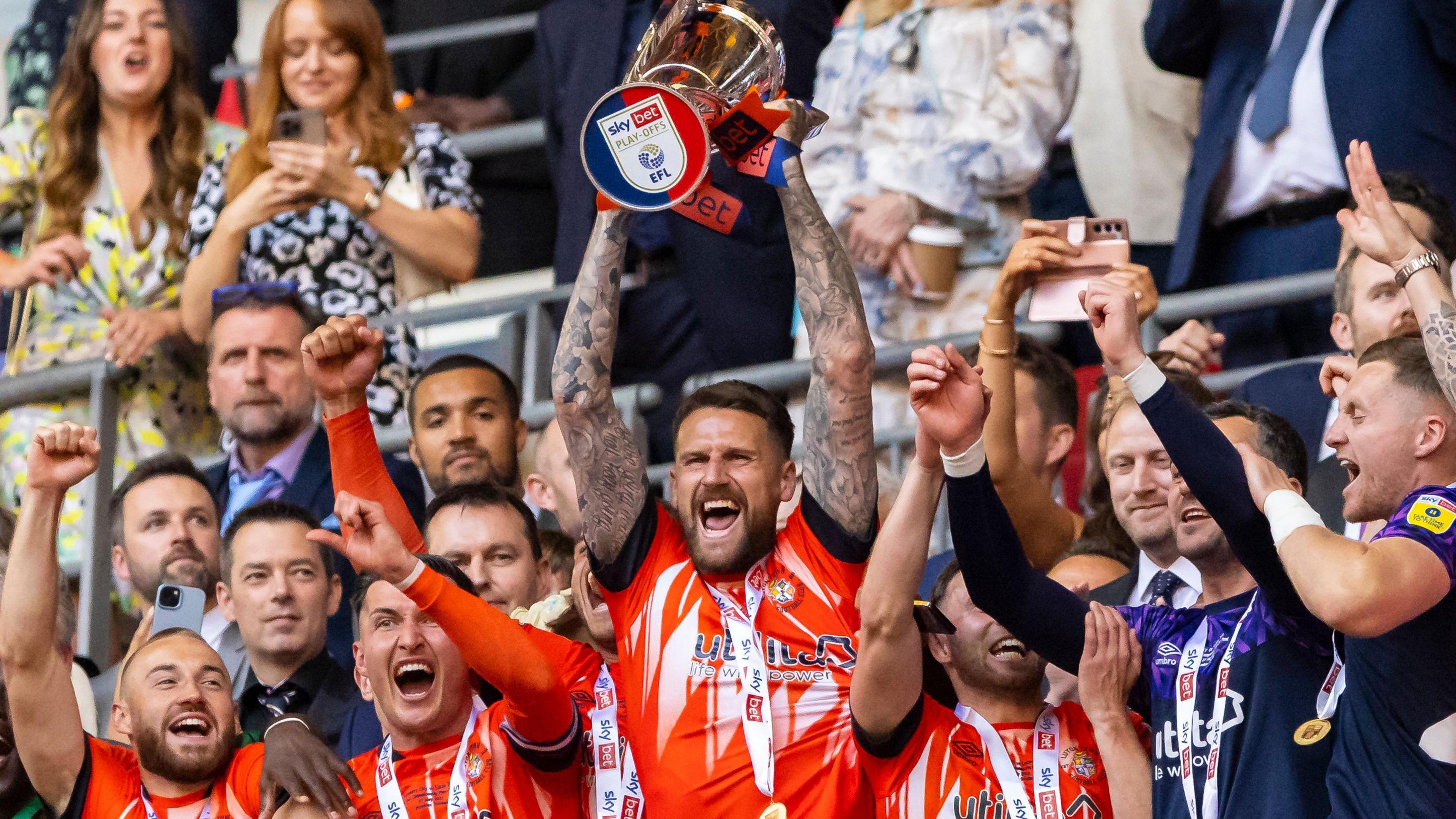 Six of the Luton Town team are visible on Wembley balcony holding EFL Trophy, wearing either orange home kit, or training tops 