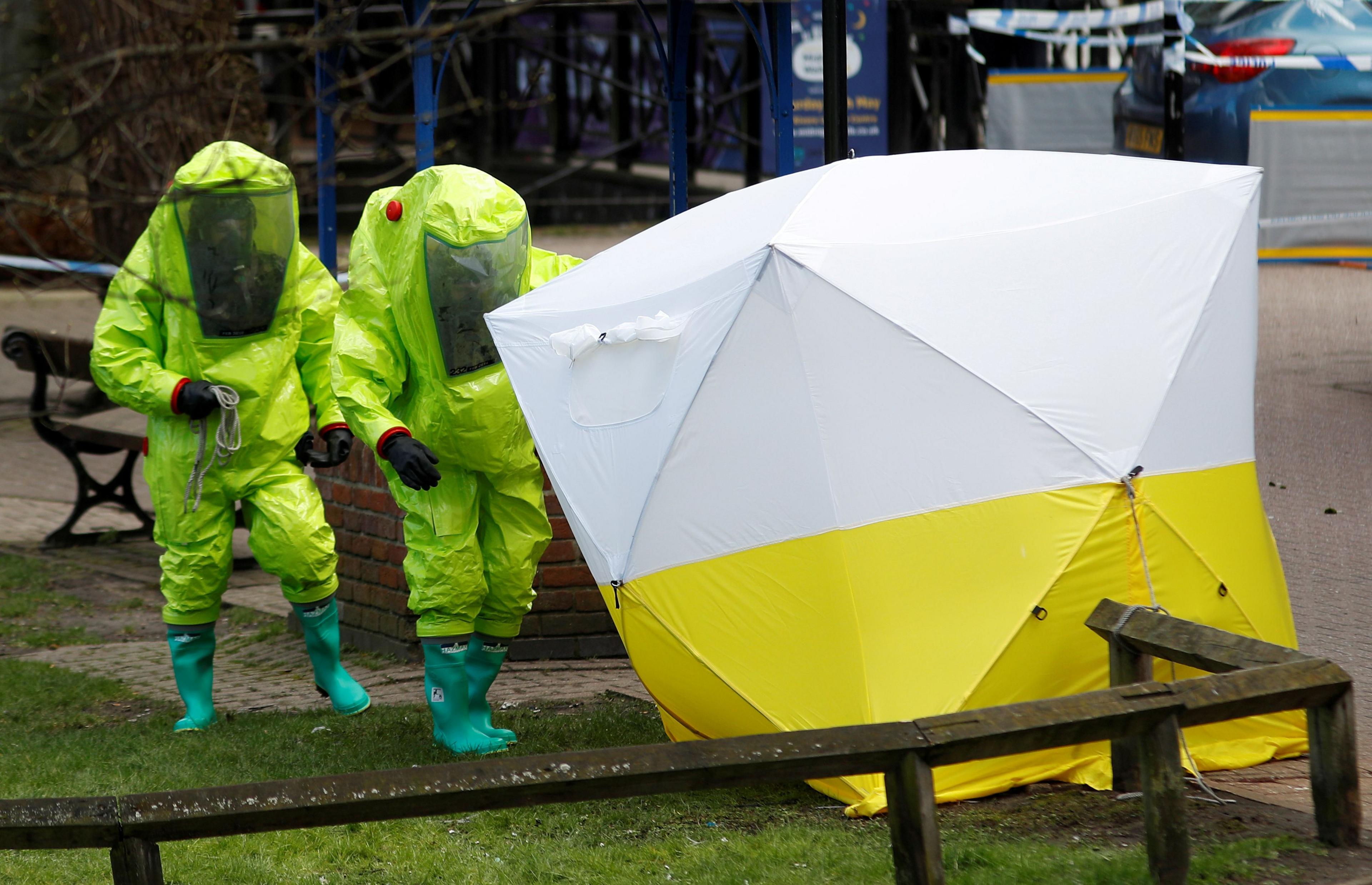 Investigators in neon green hazmat suits place a white and yellow forensic tent over a bench in Salisbury. 