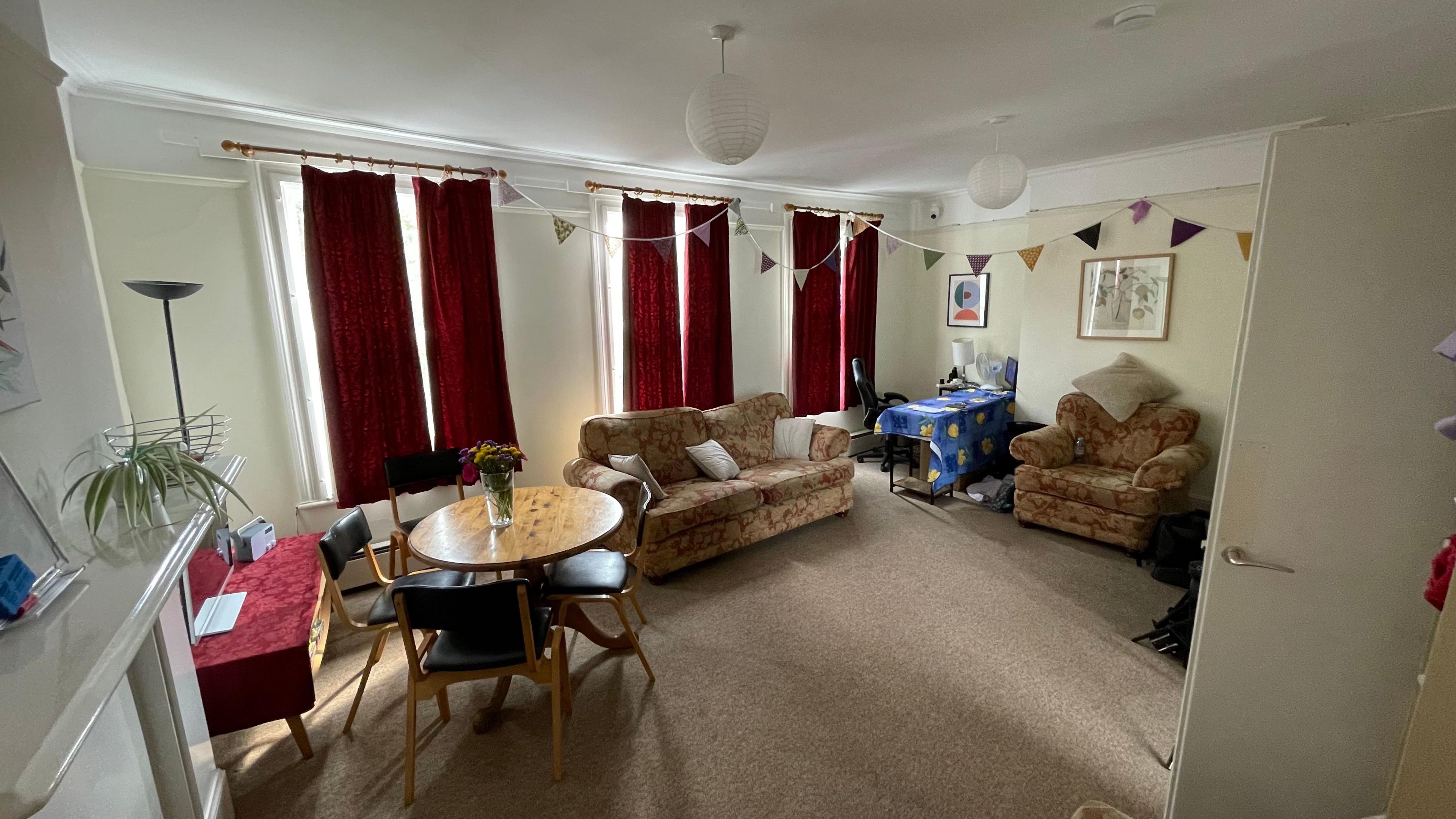 A wide angle shot of the sitting room at The Haven, taken from one corner of the room and pointing towards the opposite corner. A small, round dining table is on the left of the frame. Four wooden dining chairs with black cushioned seats and backs are arranged around the table. A two-person sofa, with a red and light brown pattern, is in the background, against one wall. The wall has three windows, which are all covered by heavy red curtains. A single-person arm chair in the same pattern as the sofa can also be seen against another wall. A small desk and office chair can just be seen in the opposite corner. There is bunting hanging between the curtain rails and picture rail.  The carpet is light-ish brown. The walls are cream coloured.