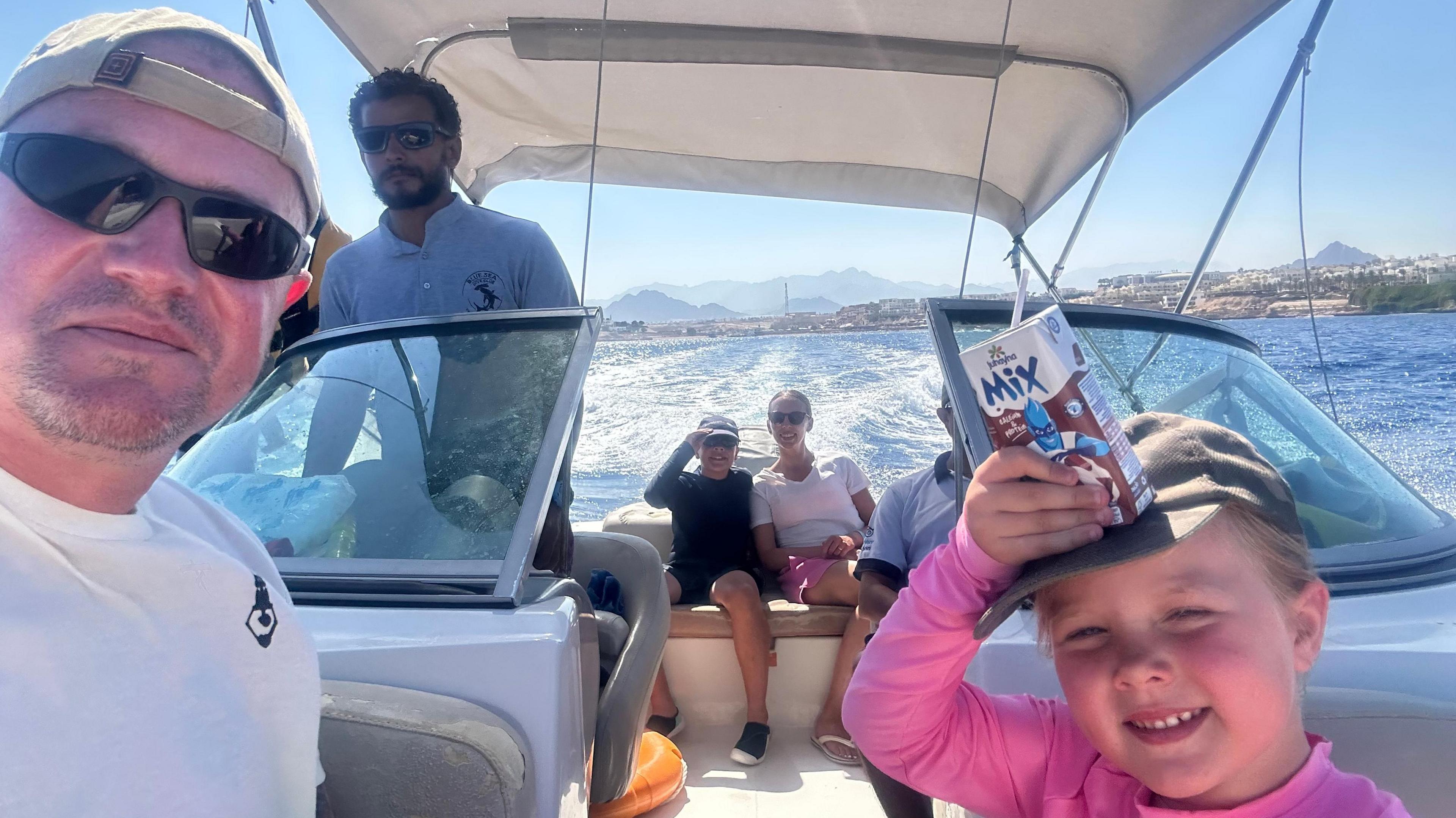 A man in sunglasses is taking a selfie on a boat with water and hills in the background, his daughter is beside him in a gap, squinting slightly and smiling, a carton of what looks like chocolate milk on her head. A boy and a woman in sunglasses are further back on the boat and a man with a T-shirt saying 'Deep Blue Dive Club' and sunglasses stands up, appearing to work on the boat.