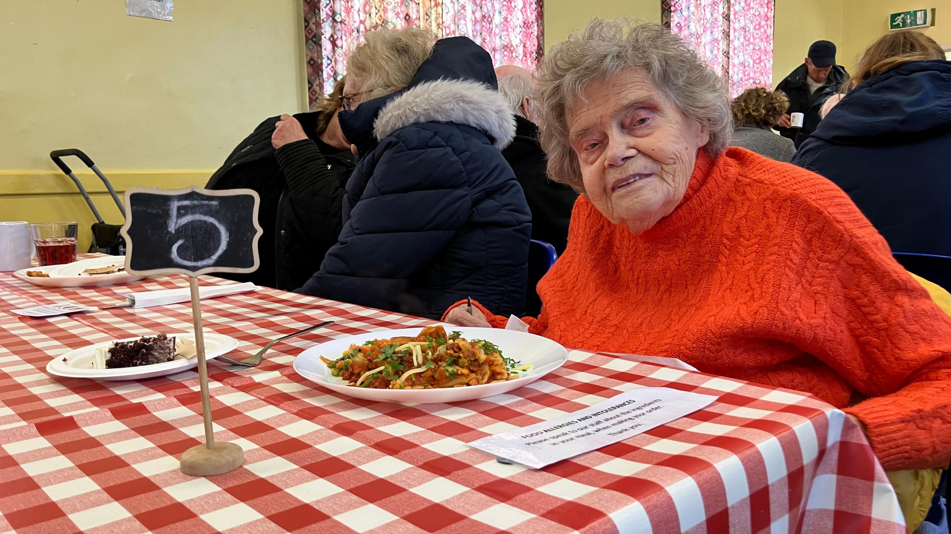 Shirley wearing a bright orange jumper looking at the camera with a plat of food in front of her 