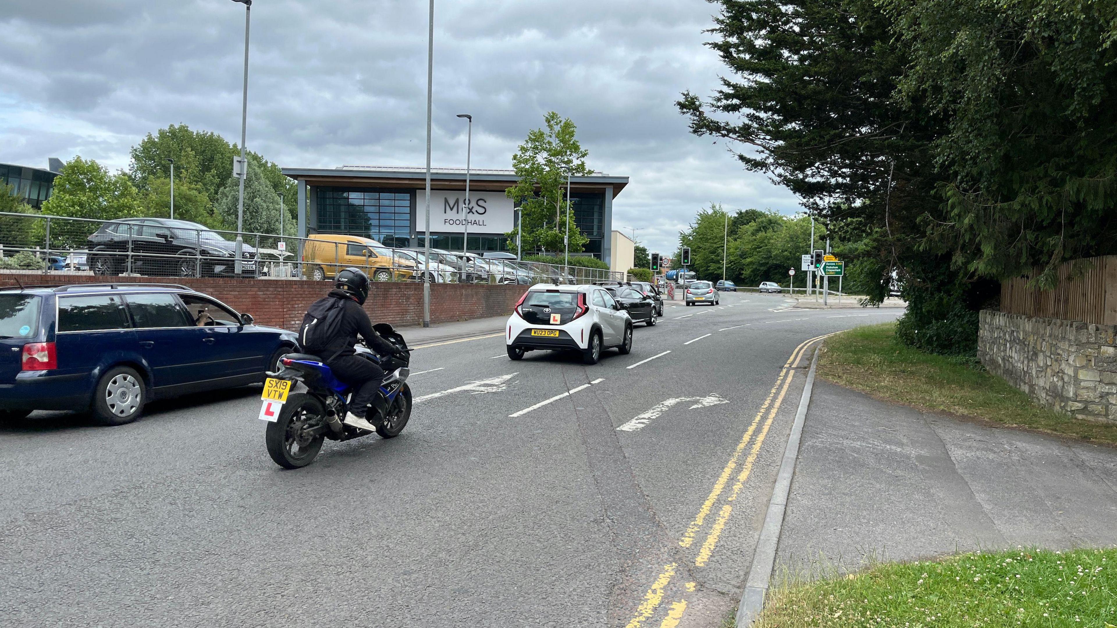 A traffic junction with several cars driving past the camera. Signage and trees are visible in the distance as is a supermarket foodhall.