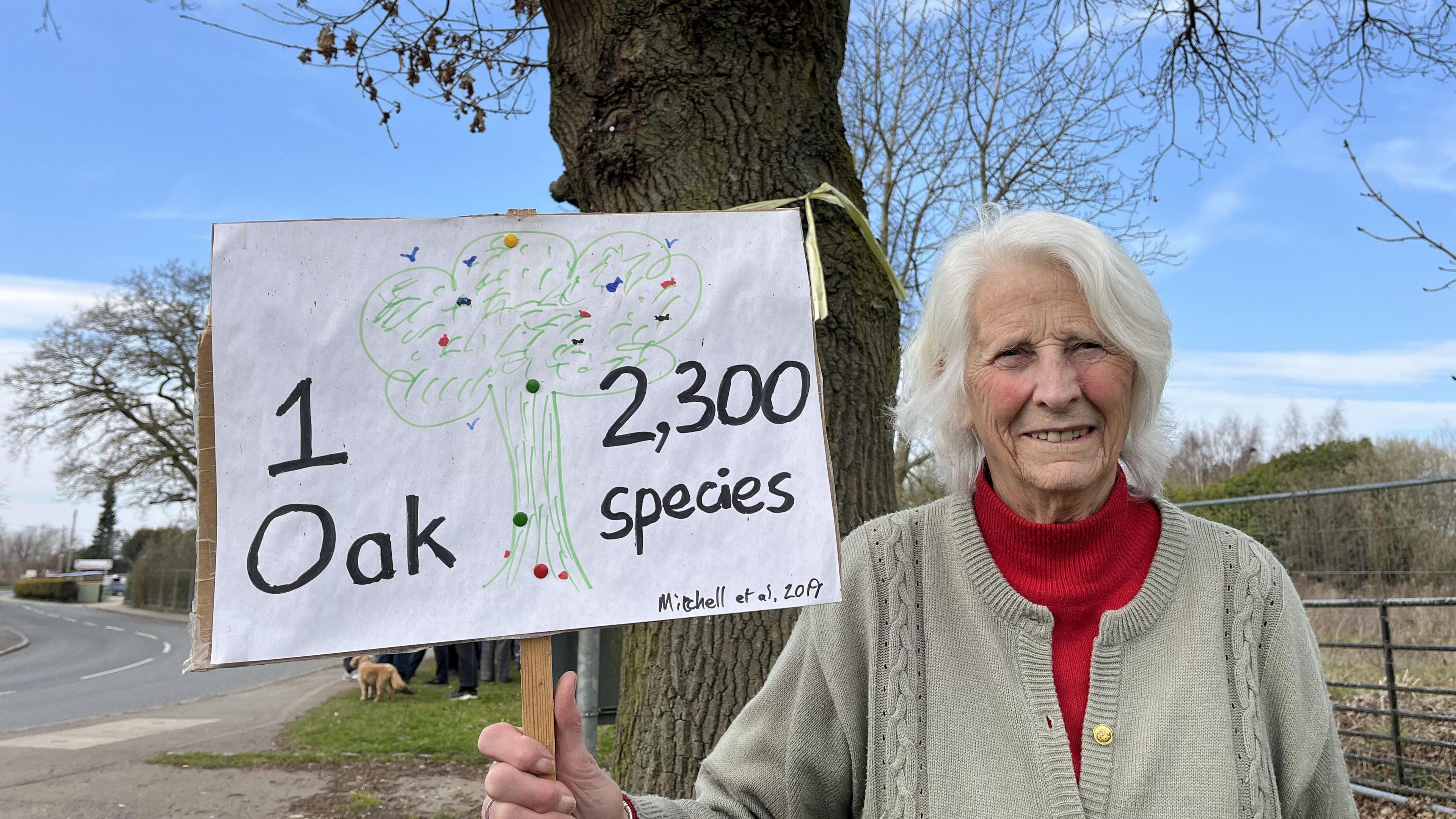 Penny stands infront of the tree holding a plaque which reads 1 oak, 2,300 species.   