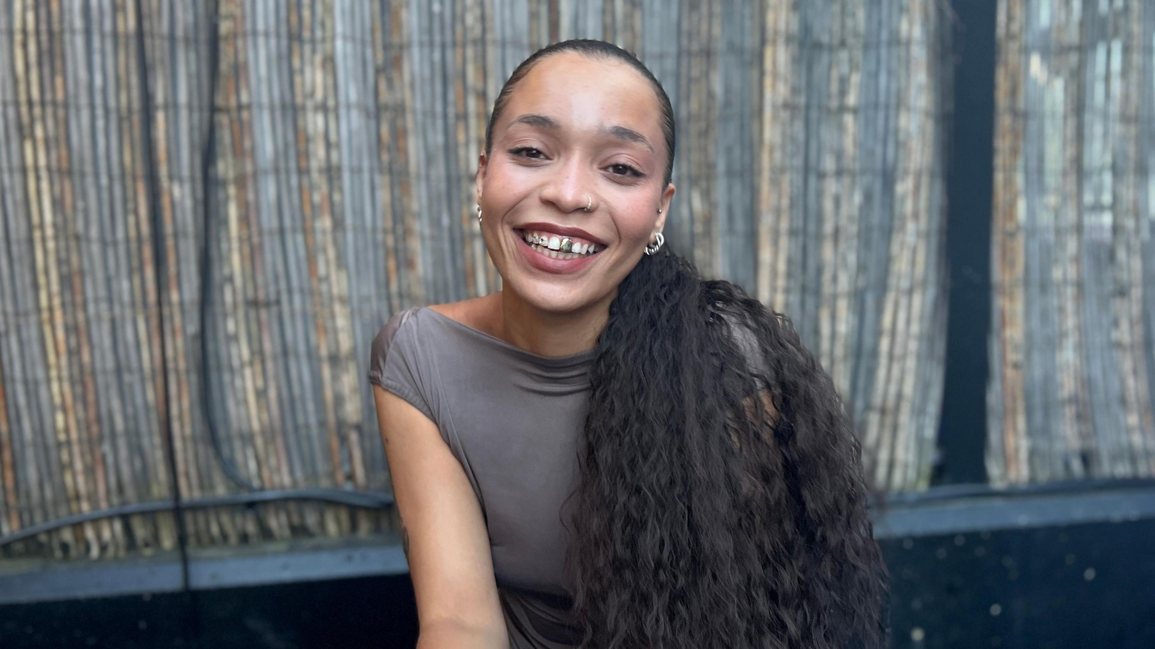 A woman with long black hair tied back into a loose ponytail slung over her left shoulder sits in front of a bamboo screen, smiling. She's got a silver front tooth and several small silver hoops in each ear.