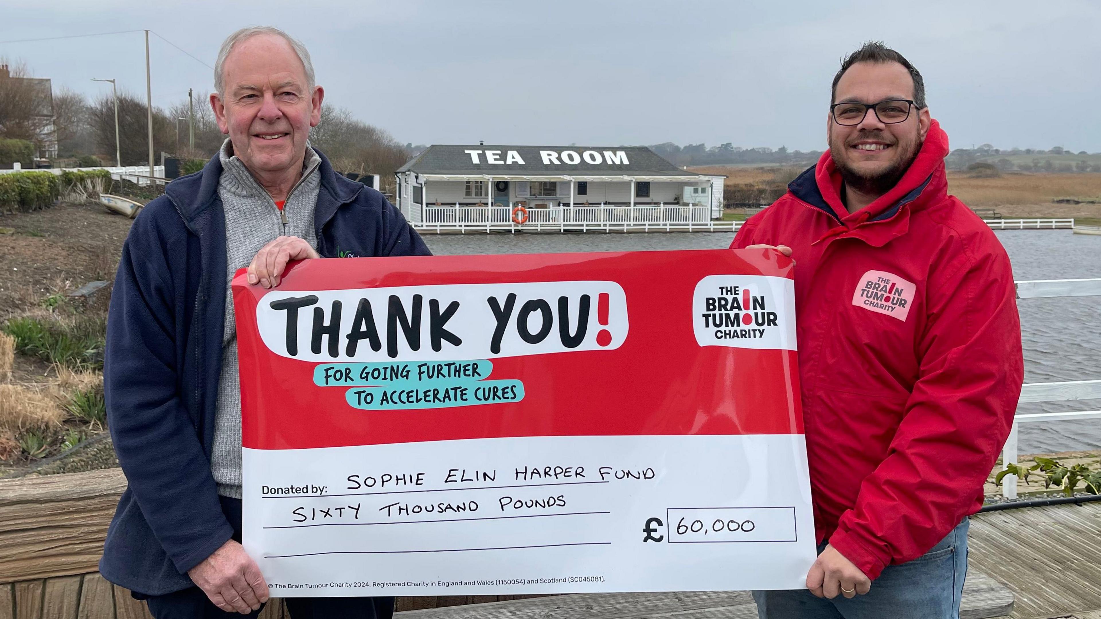 Mr Huggins, dressed in a navy fleece and grey zip-up pullover, holds a giant cheque for £60,000 donated by the Sophie Elin Harper Fund. The cheque is also branded with the words: thank you for going further to accelerate cures next to The Brain Tumour Charity logo. A man wearing a red charity coat holds the other side of the cheque. The pair stand in front of Southwold boating lake and tearoom.