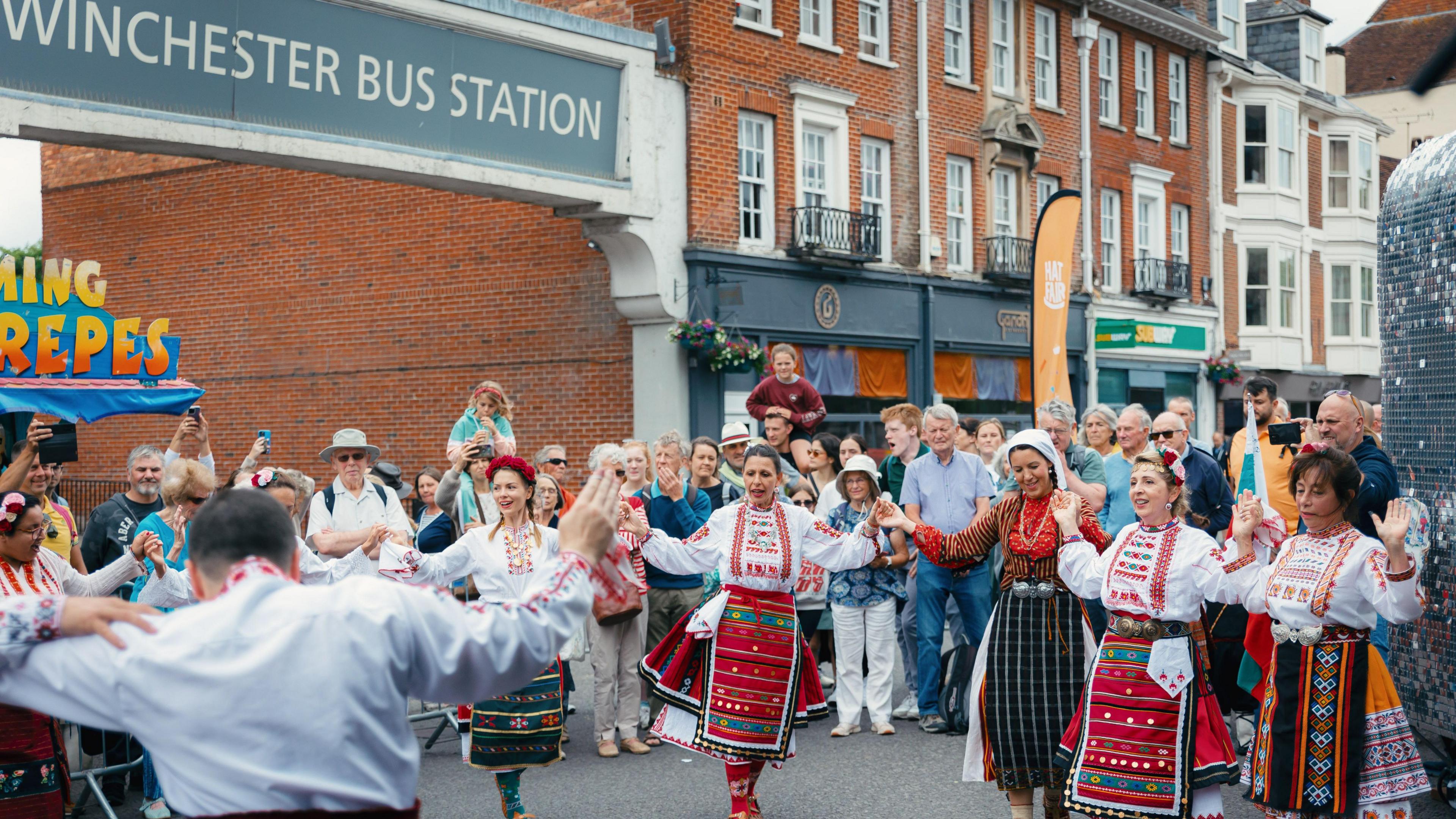 A group of women in traditional costumes dance in the street outside Winchester Bus Station with a large crowd watching on.