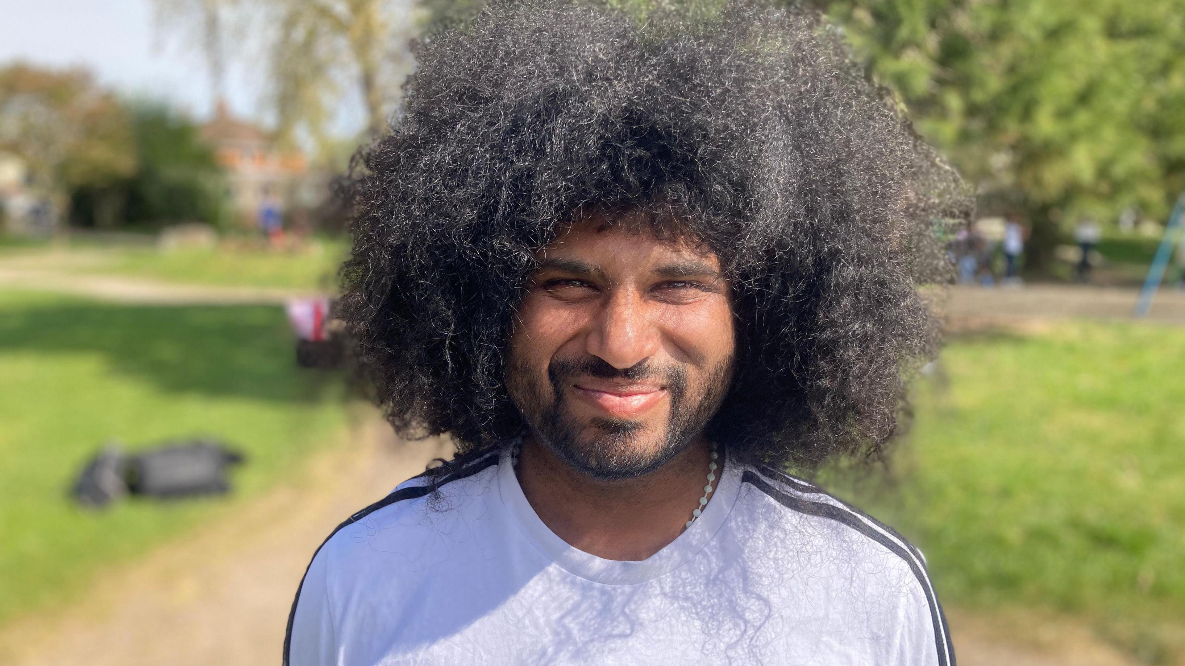 Vance Gregory Machado pictured smiling slightly at the camera, with the blurred background of a park behind him. He has long black curly hair and is wearing a grey t-shirt with black stripes on the shoulders.