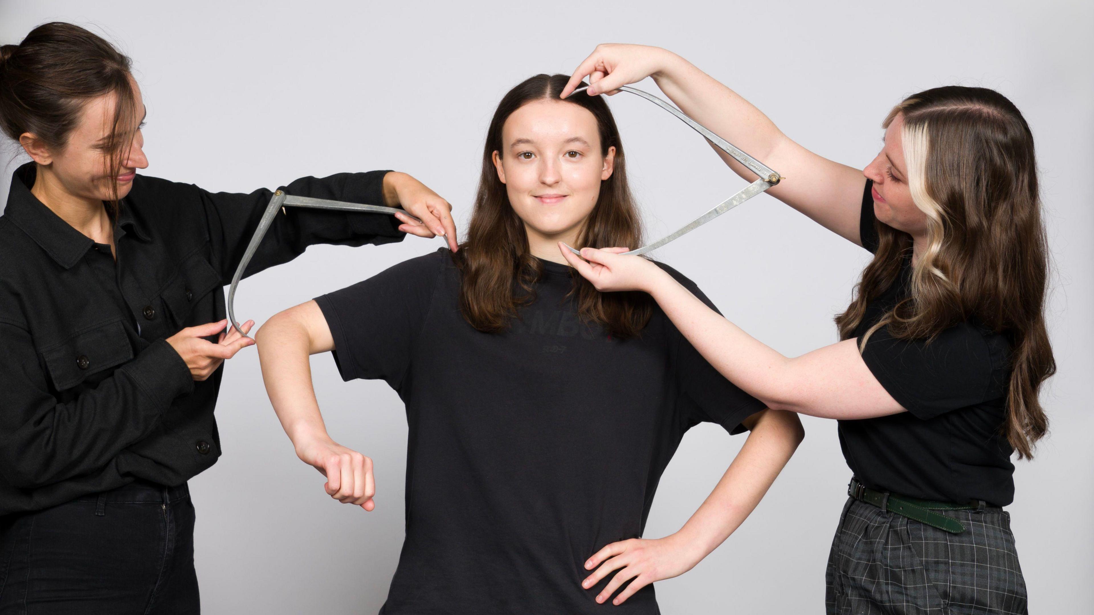 Bella Ramsey during the measuring up process for their first figure for Madame Tussauds London. Two people are either side holding measuring equipment. All three are dressed in black.
